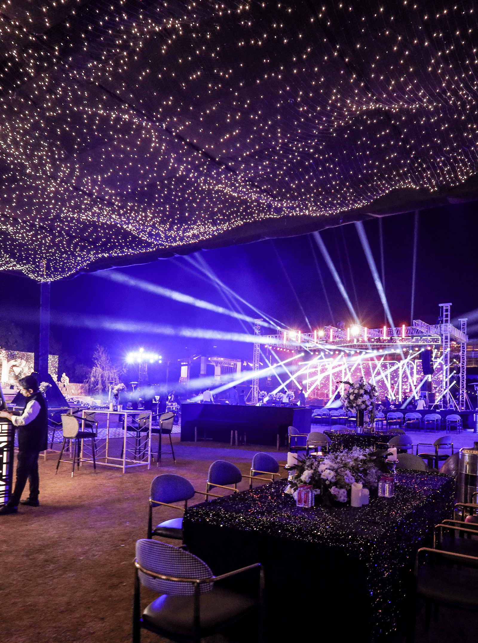 An outdoor event at Ananta Spa & Resort, Jaipur, features a stage with blue lighting and a striking canopy of string lights.