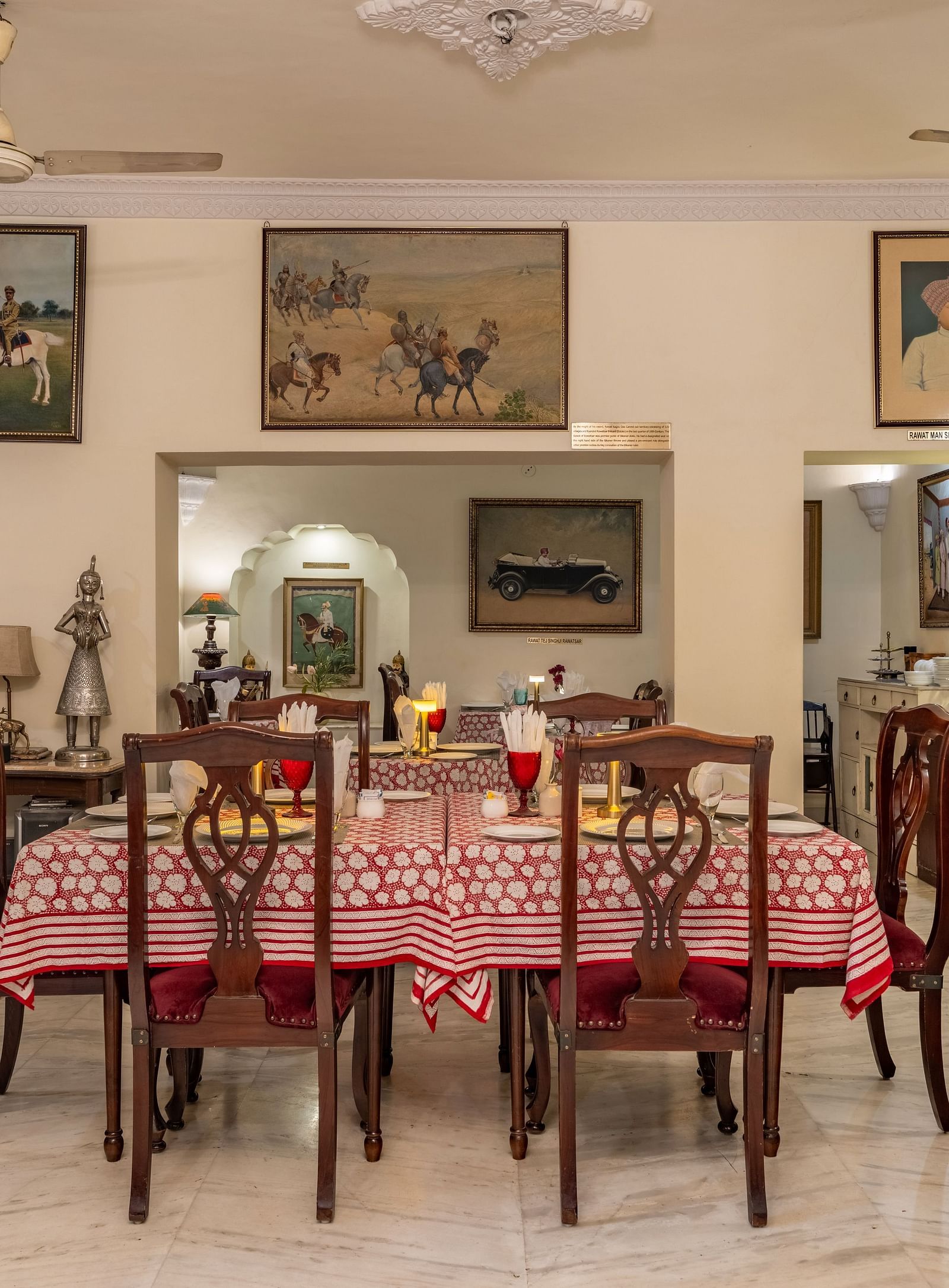 A dining room with set tables, wooden chairs, artwork on the walls, ceiling fans, and a glimpse into another room | Dera Rawatsar, Jaipur