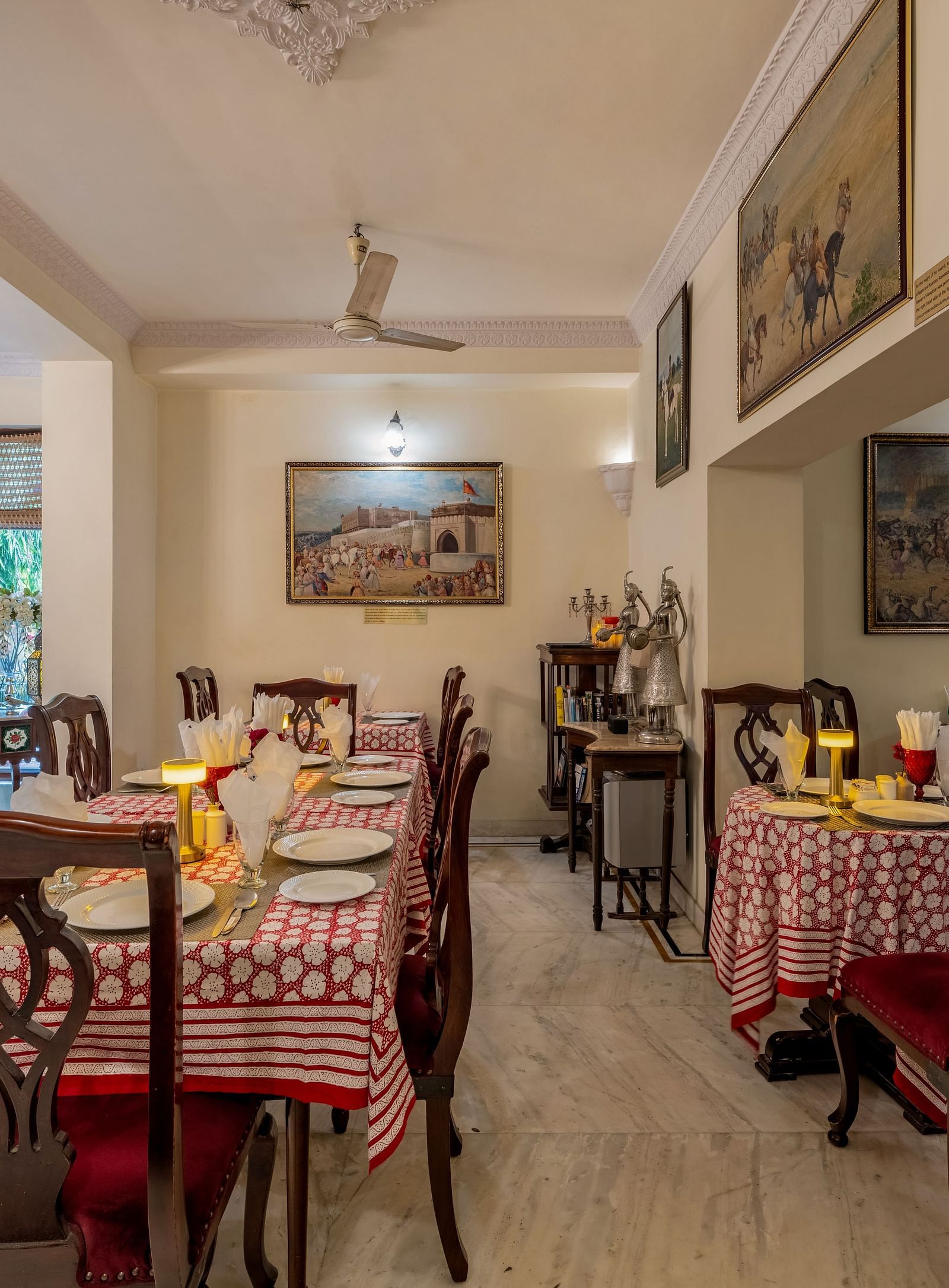 A long, narrow dining room with set tables, wooden chairs, artwork on the walls, and ceiling fans | Dera Rawatsar, Jaipur