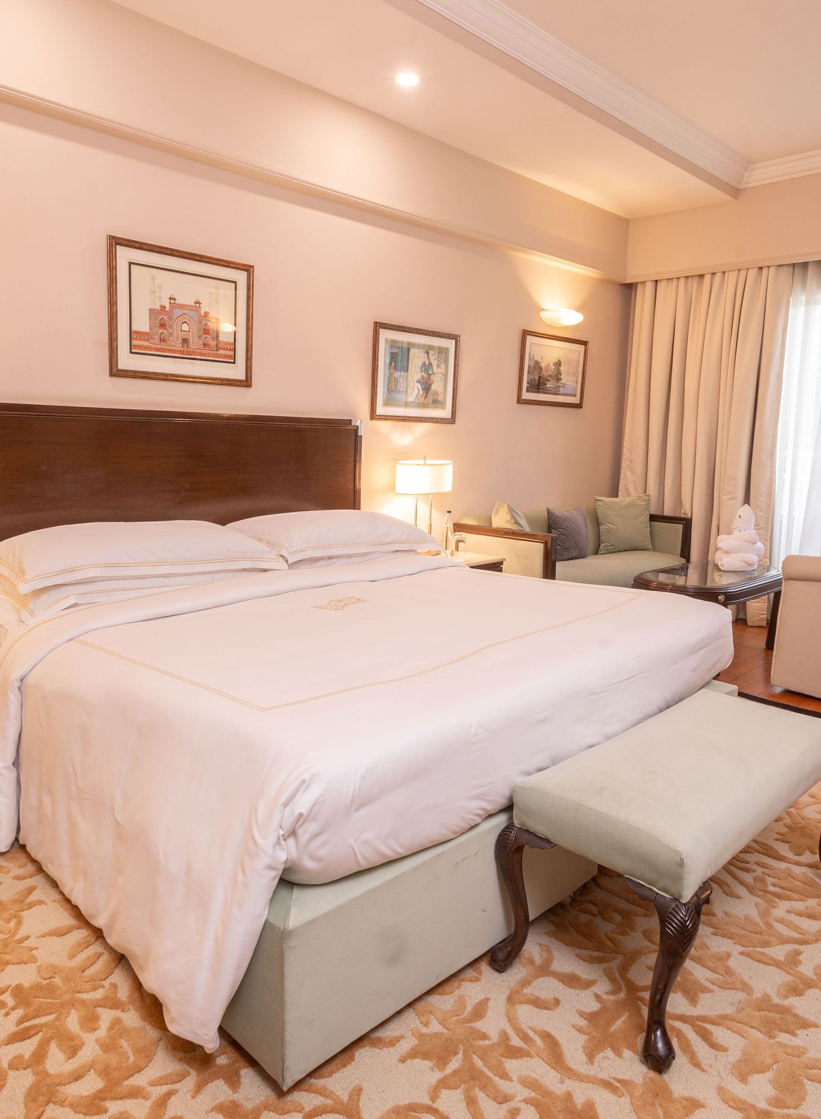 side angle view of the heritage balcony room with a plush comfortable king size bed and a cushioned stool in front of it - The Claridges New Delhi