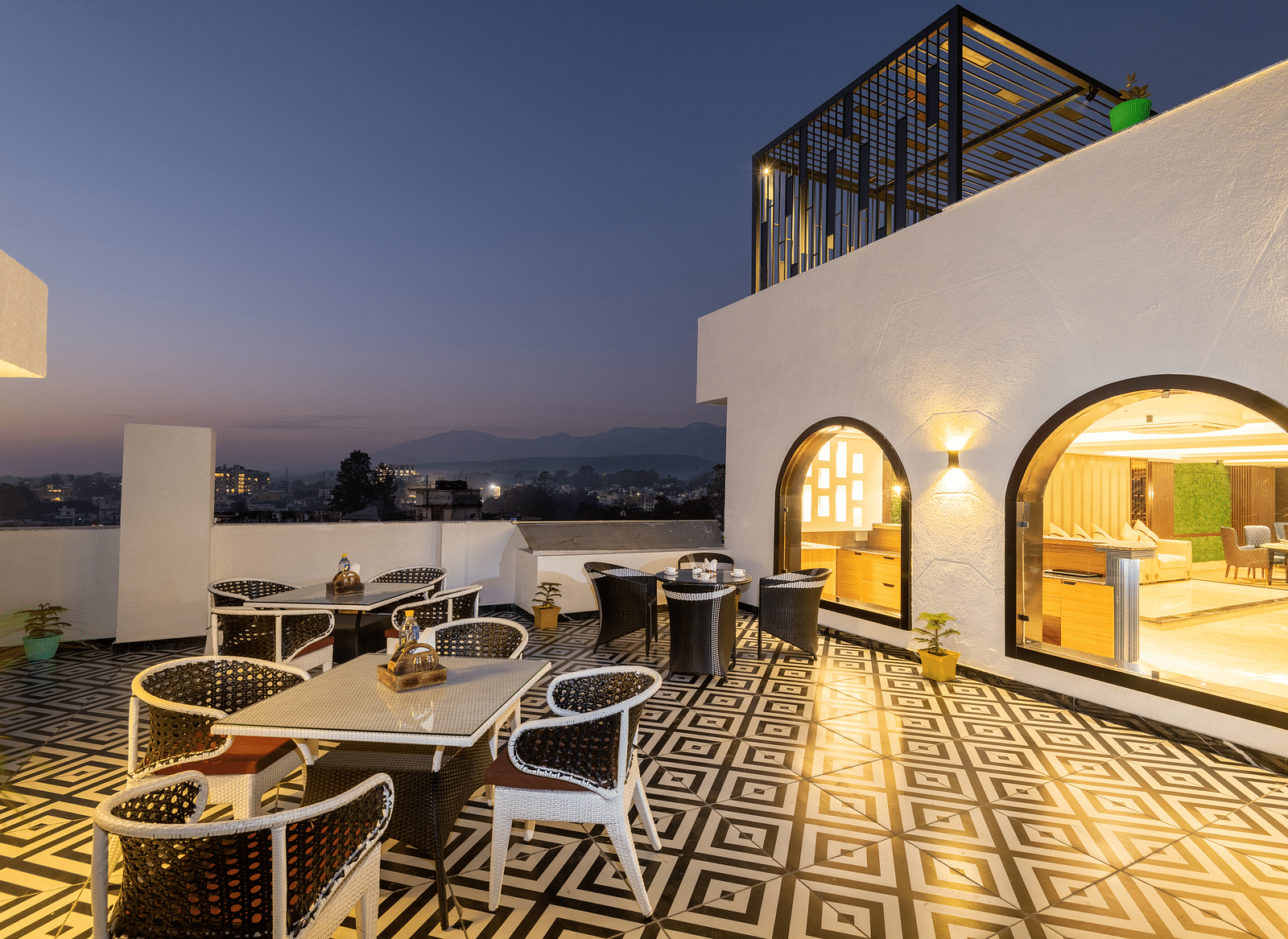 Image of the rooftop restaurant at DLS Grand Luxotica, Dehradun during evening featuring chairs and tables arranged on a well-furnished floor