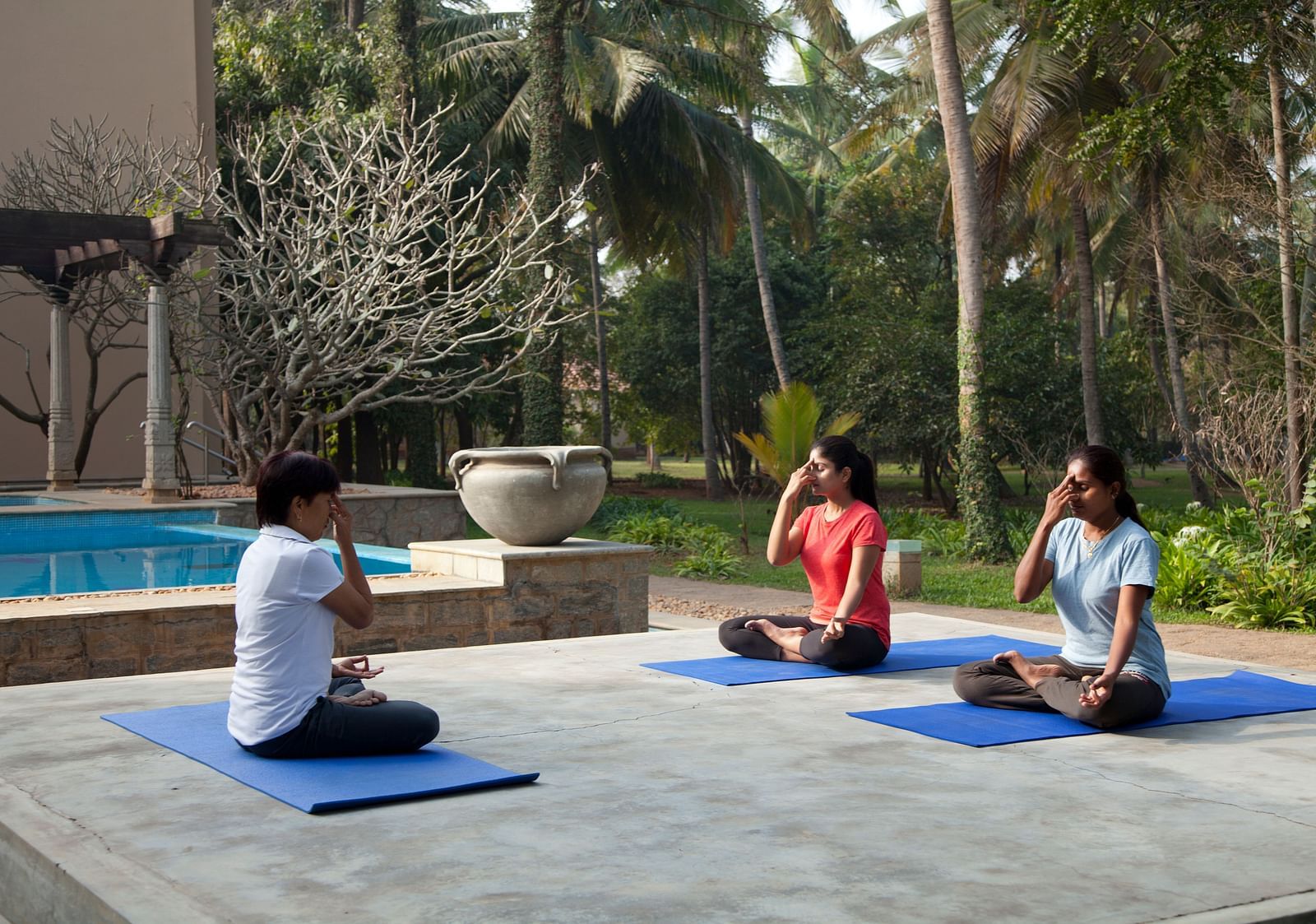 People doing yoga poses outdoors by a pool, trees in background - Shreyas Retreat