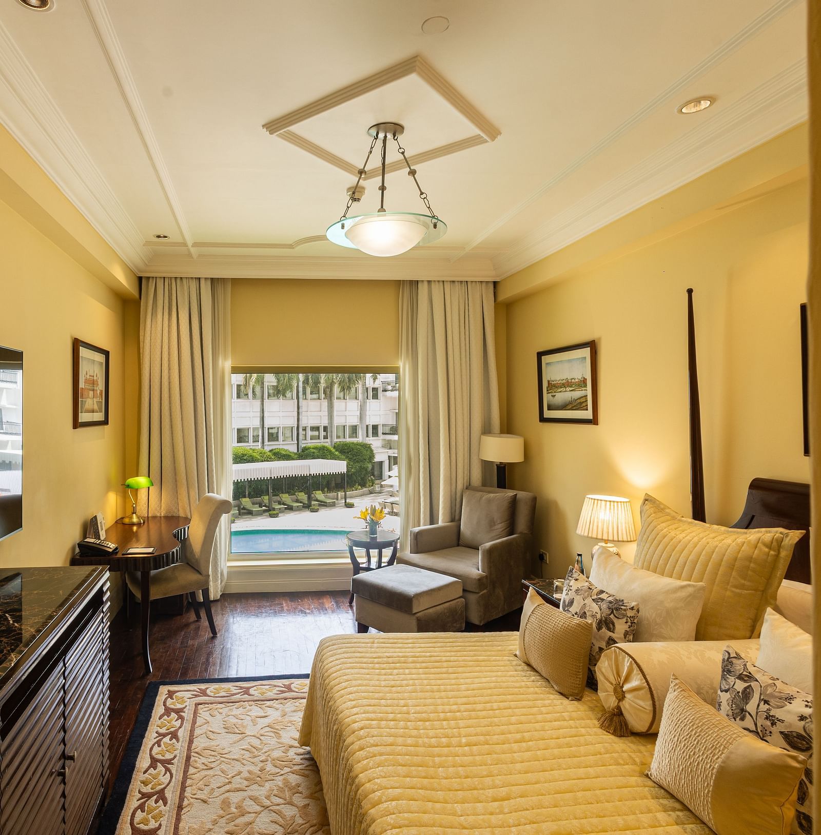 Side view of the Deluxe King Bed Room Pool View at The Claridges New Delhi, featuring a cosy king sized bed, wall-mounted TV, a corner chair, and a large window overlooking the swimming pool
