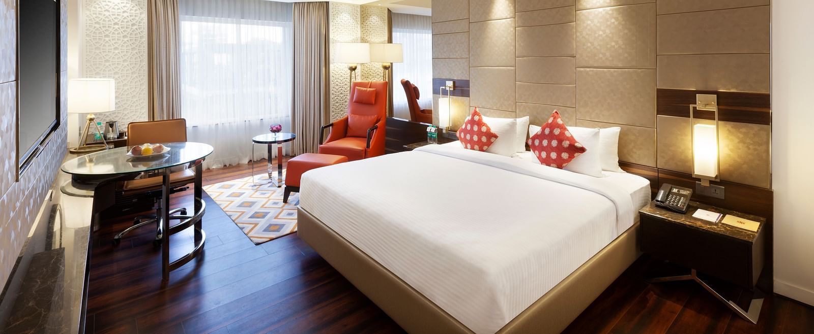 A spacious room with a large bed, wall-mounted TV, table, and cushioned chair, with curtains pulled to the side of the windows | Cenneys Gateway