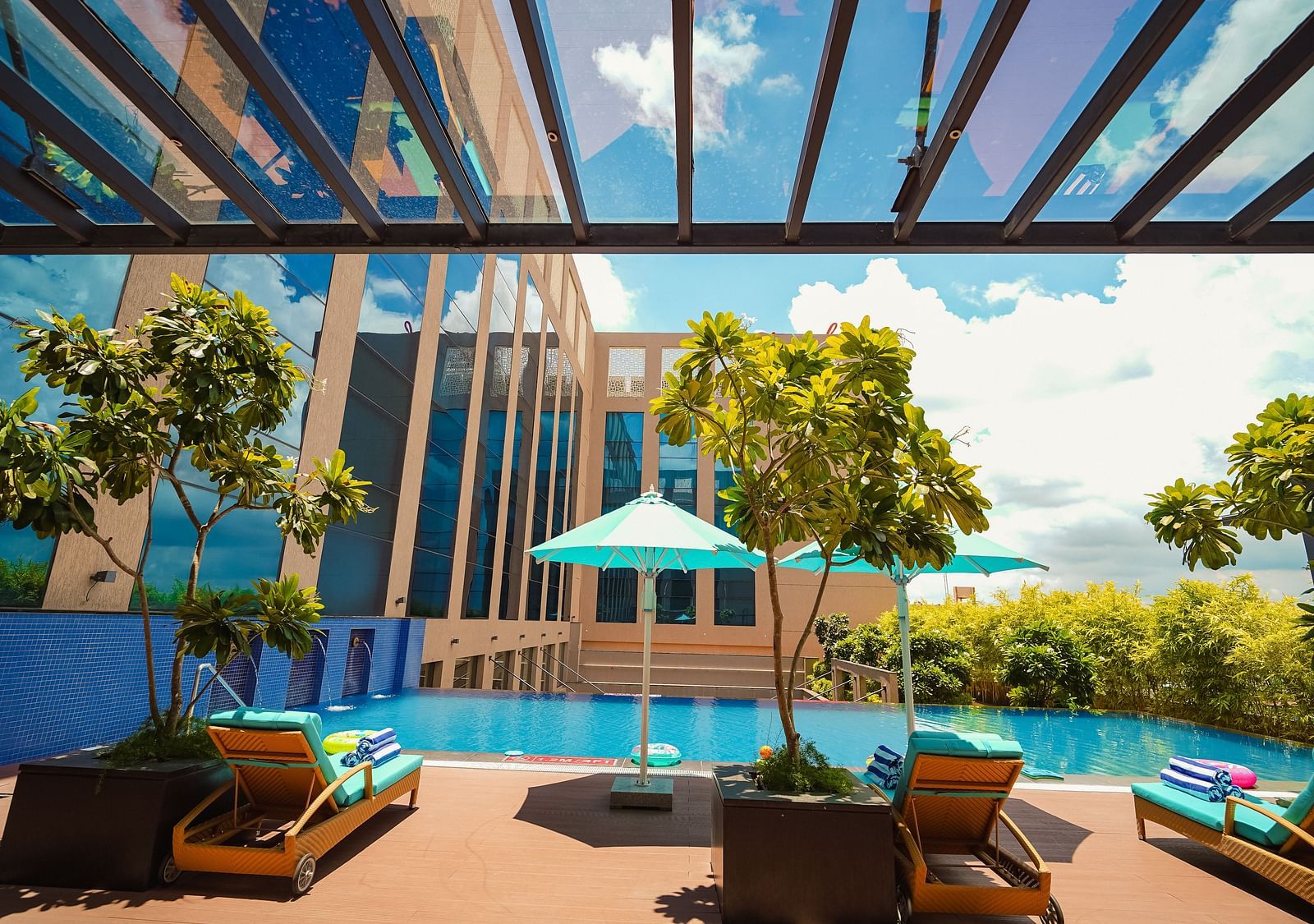 A serene view of the pool, reflecting the blue skies, surrounded by lush greenery and shaded seating areas - Grand Madurai by GRT Hotels