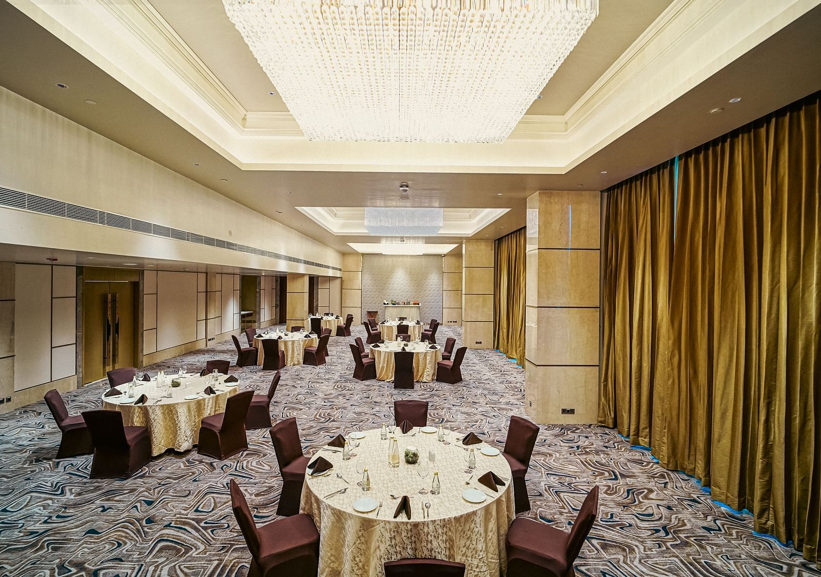 A banquet setup in a large hall with round tables, cushioned chairs, and stylish chandeliers