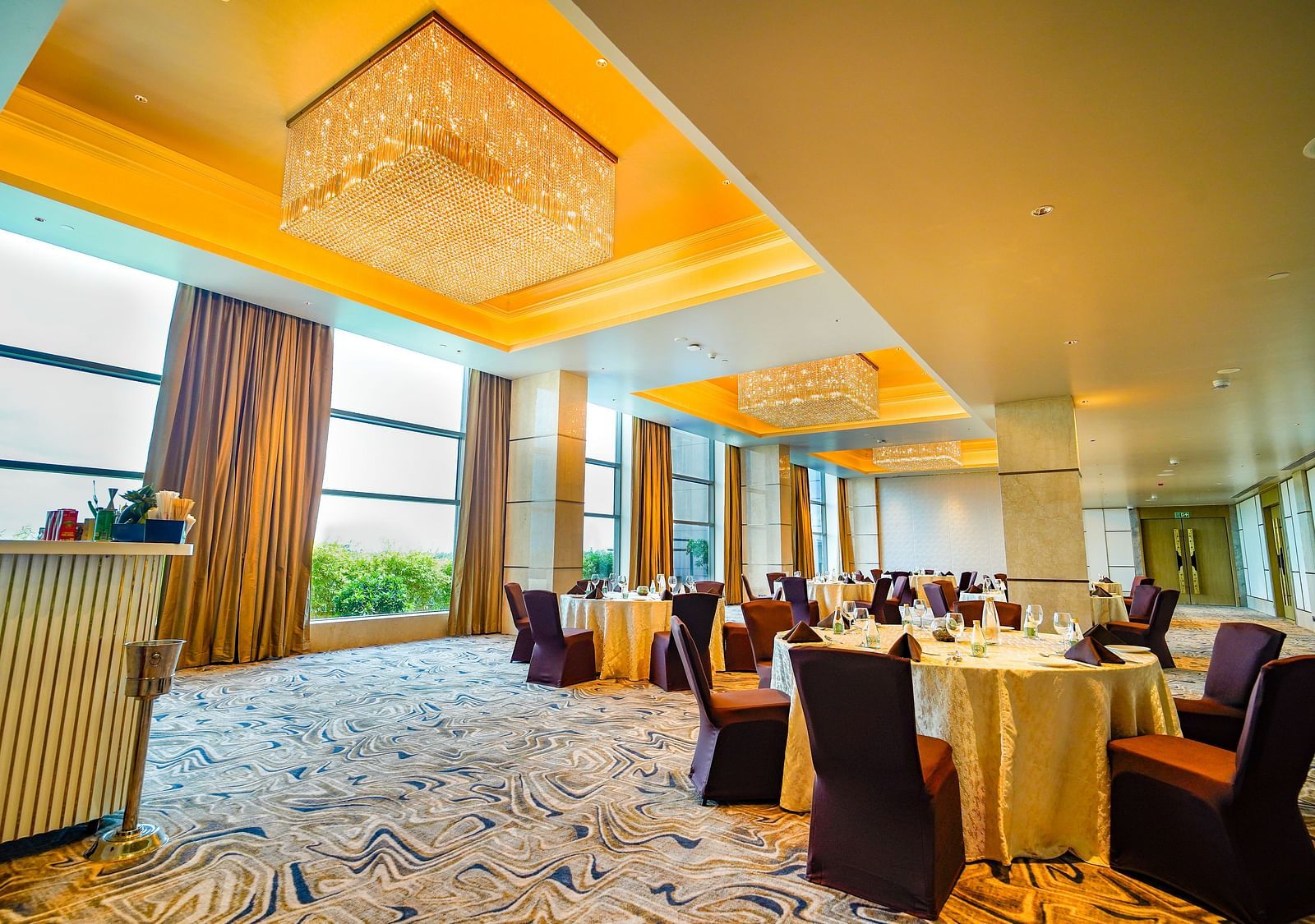 A spacious banquet hall with round tables, elegant chandeliers, and natural light from large windows