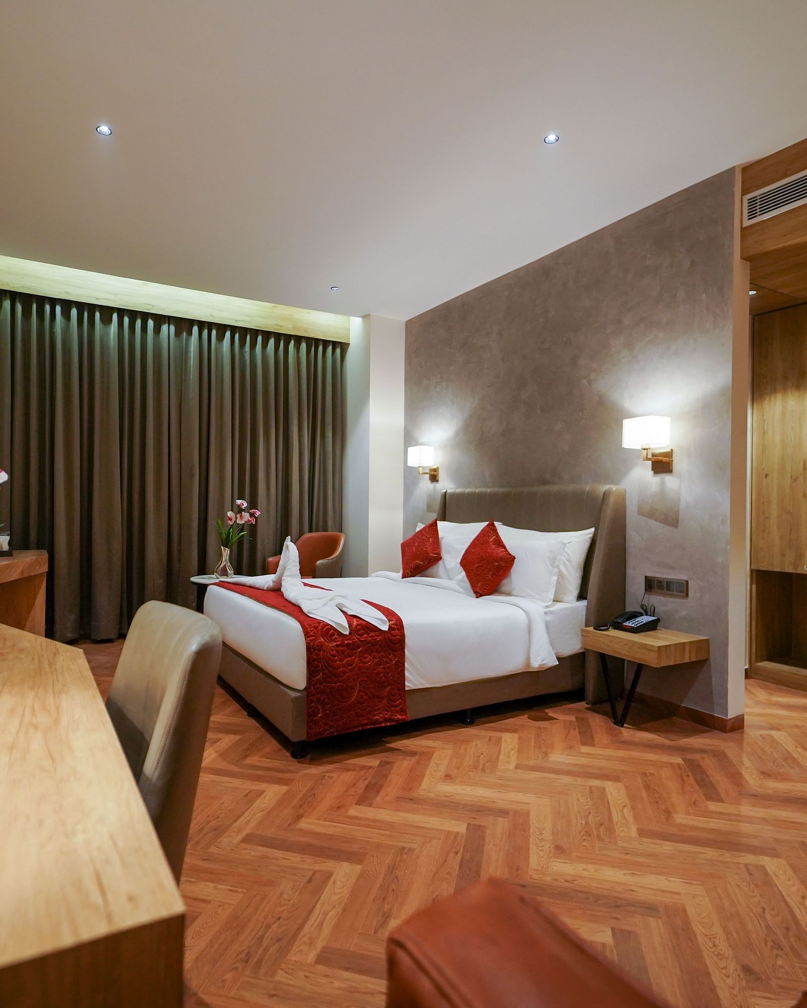 A hotel room displays a bed with red accents, a desk with a lamp, and a chair on a patterned floor - Hotel Park Inn, Nellore.