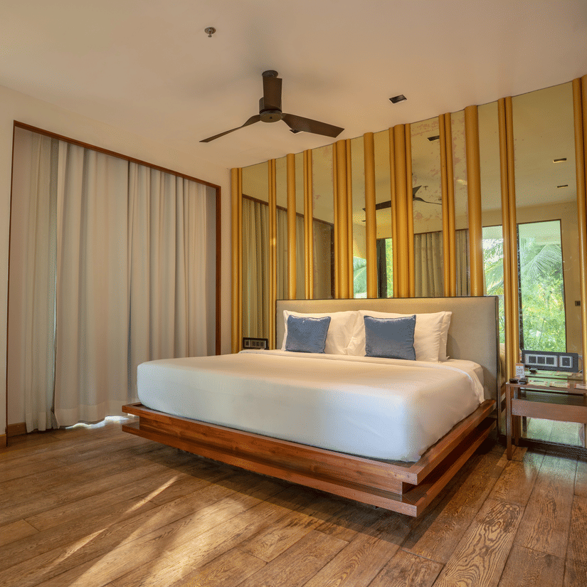 A bright and airy hotel bedroom with a large bed, wooden floors, and a private balcony. The decor features light-colored wood and a ceiling fan.