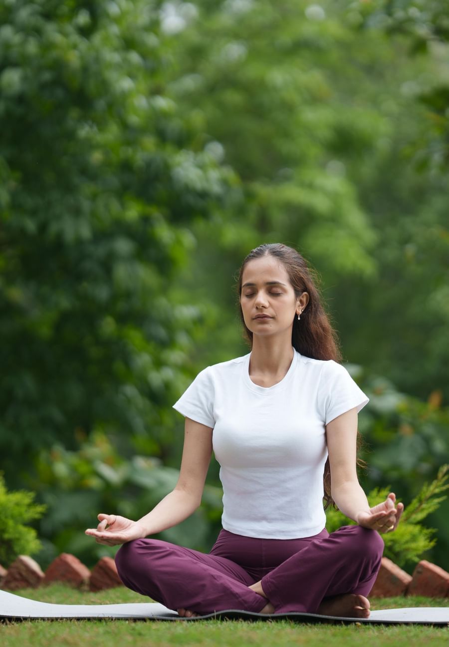 A woman in a white t-shirt and purple yoga pants sits in a meditative pose on a yoga mat outdoors amidst lush green trees, likely at  Summit Enigma Resort and Spa.