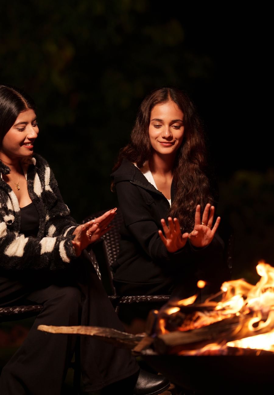 Two women are gathered around a campfire at night, smiling and enjoying the warmth, a common evening activity at Summit Enigma Resort and Spa.