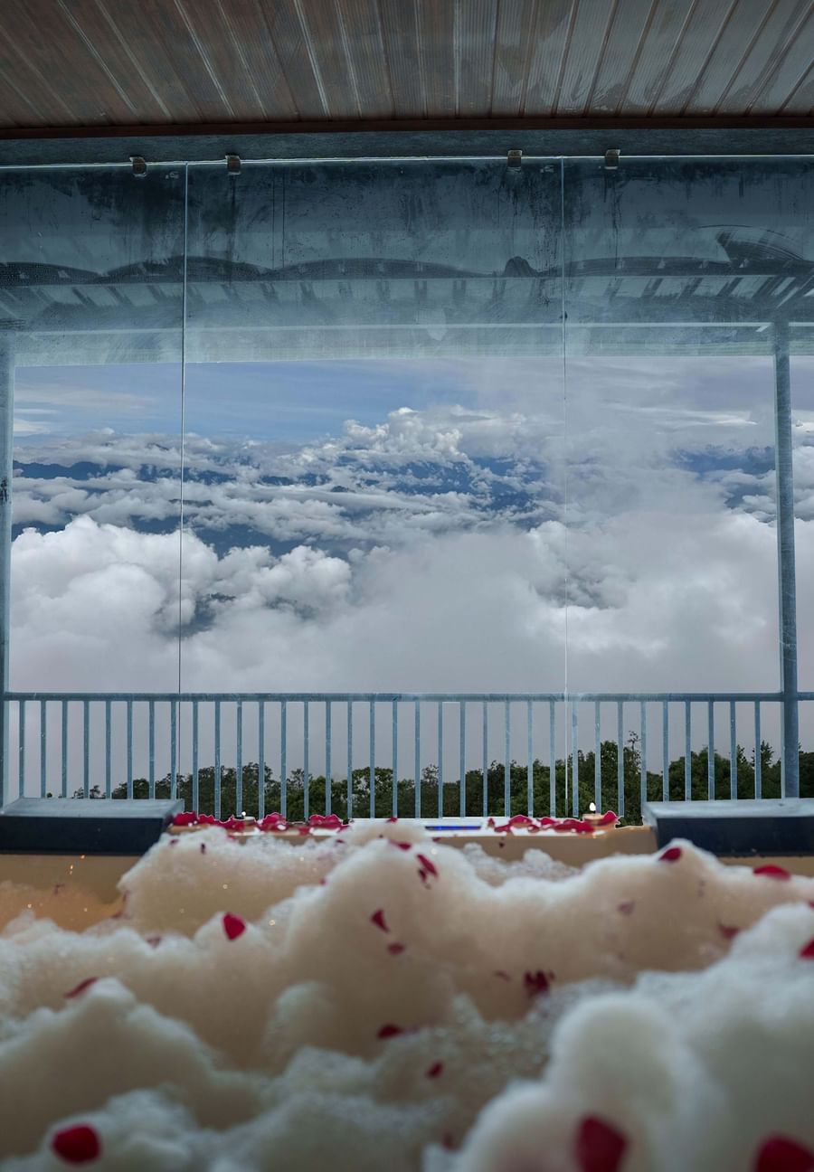 A jacuzzi with rose petals scattered, leading to a balcony overlooking a cloudy sky and distant hills, suggesting a romantic setup at Summit Enigma Resort and Spa.