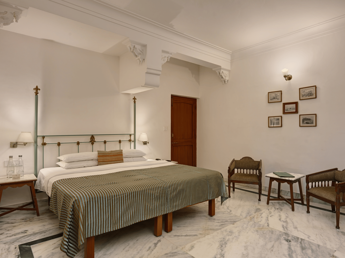 A  bedroom at Ram Pratap Palace, Udaipur, featuring a well made four-poster bed, two wooden chairs, a small table, and framed art on the wall.