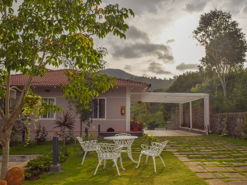 A white house with a red roof, white outdoor table and chairs on a green lawn, surrounded by trees under a bright sky.