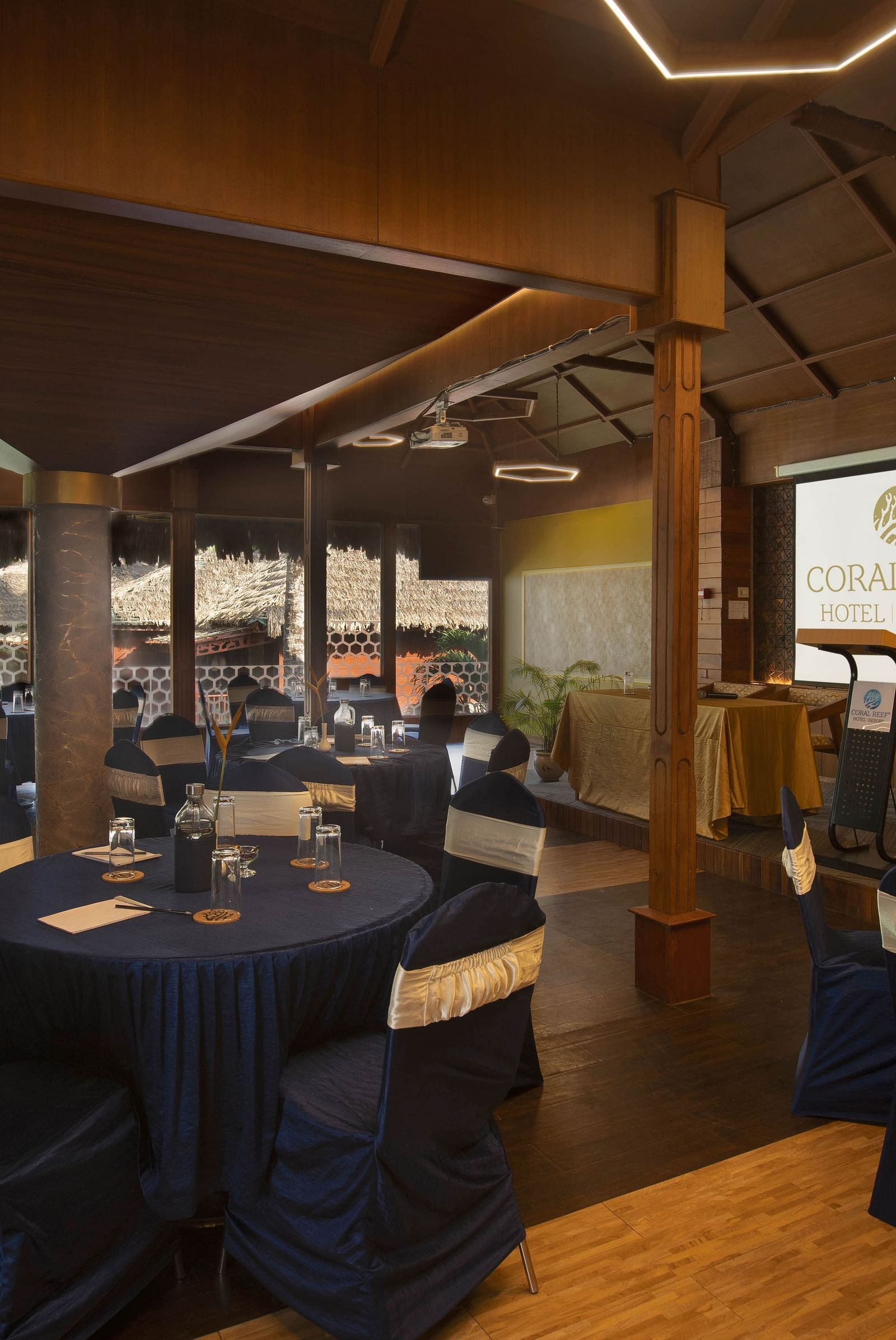 A modern, elegantly set-up banquet hall featuring dark blue table cloths and chairs, with a screen in the background for presentations. The room is well-lit with contemporary lighting - Coral Reef Resort & Spa, Havelock