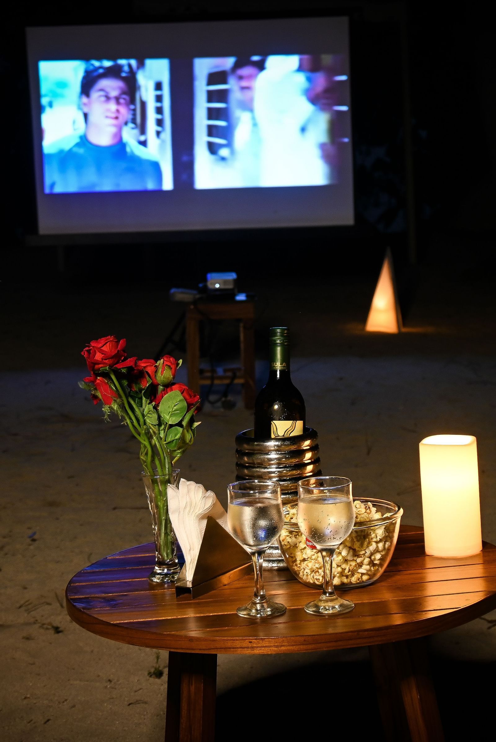 A small table with glasses wine rose on top and a movie playing in the white screen in the background - Coral Reef Resort & Spa, Havelock