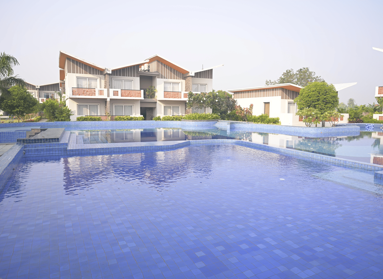 A calm swimming pool reflecting nearby villas and greenery on a sunny day at Hotel Sonar Bangla Mayapur.