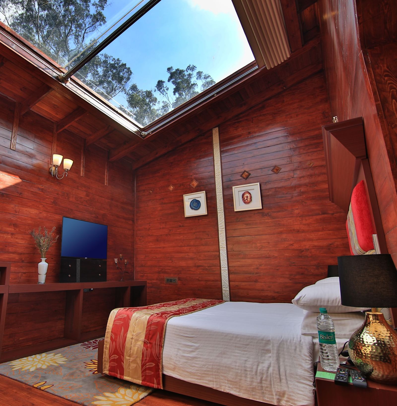 A wide angle image of a room in Ooty featuring a bed, a television and a glass roof flooding the space with sun light - Accord Highland Ooty