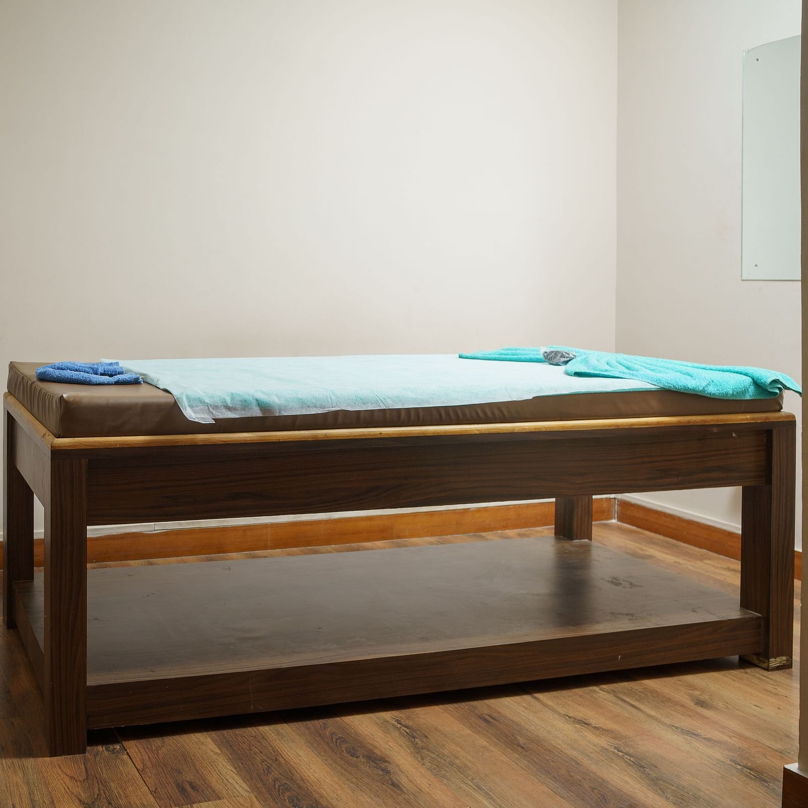 A wooden massage table in a room with light-coloured wooden flooring and white walls, with some folded towels on a shelf at Orsino Spa Resort.