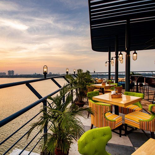 A scenic rooftop restaurant with colorful chairs, wooden tables, and a view of the sunset over a river. - Polo Floatel