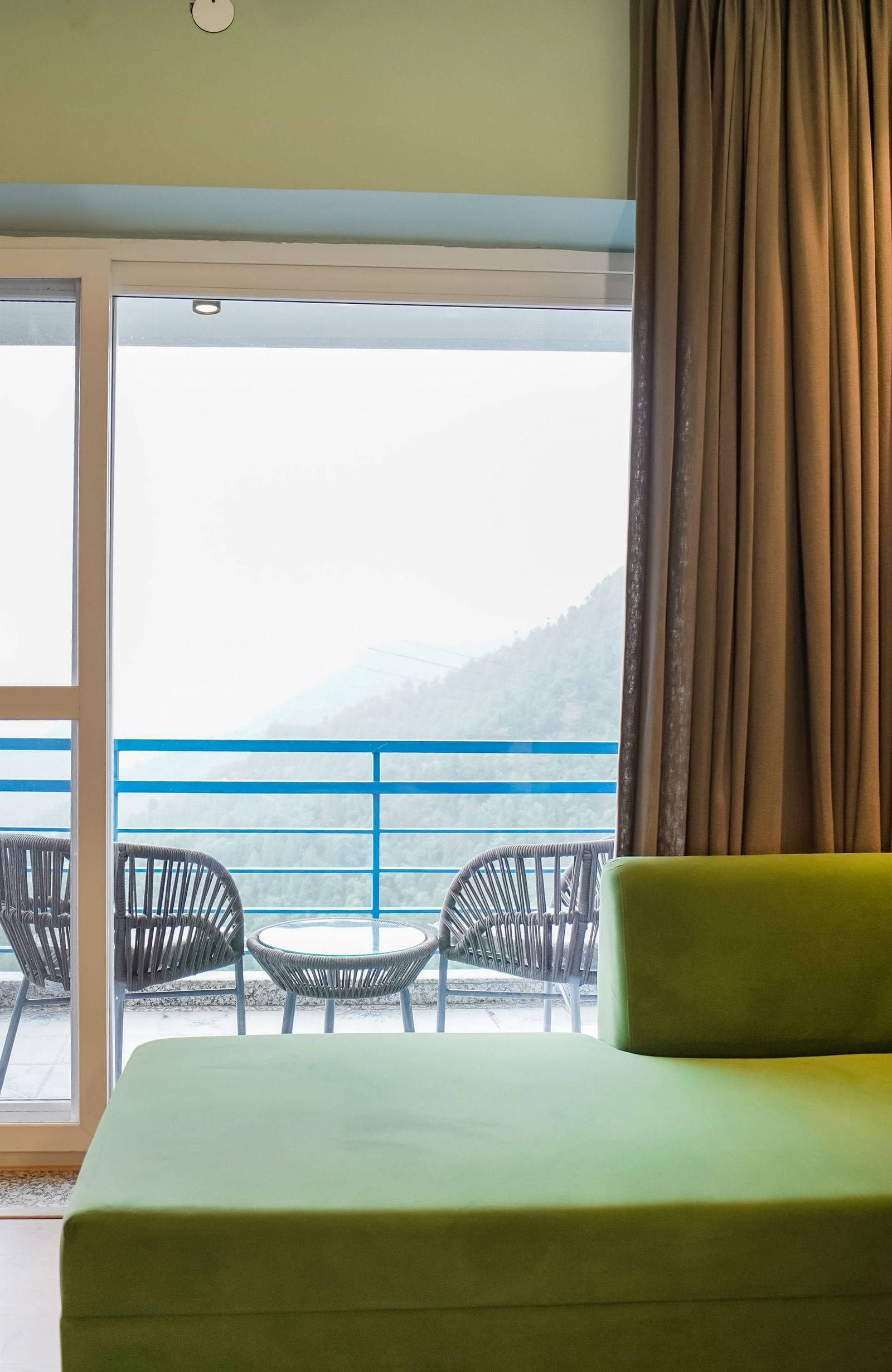 A modern hotel room with a green sofa and a large sliding glass door opening to a balcony with outdoor seating and a view of the hills | Deluxe Room with Valley View | Sukama Resort & Spa, Nainital