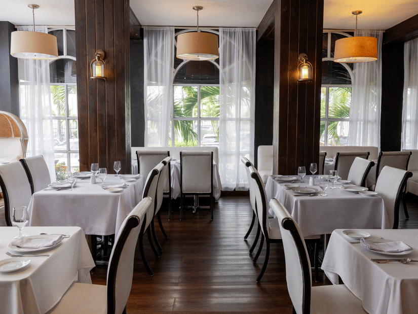 The dining area of Rocksteady Restaurant with rows of tables covered with tablecloths, cushioned chairs, pendant lights, and large windows at S Hotel Kingston.