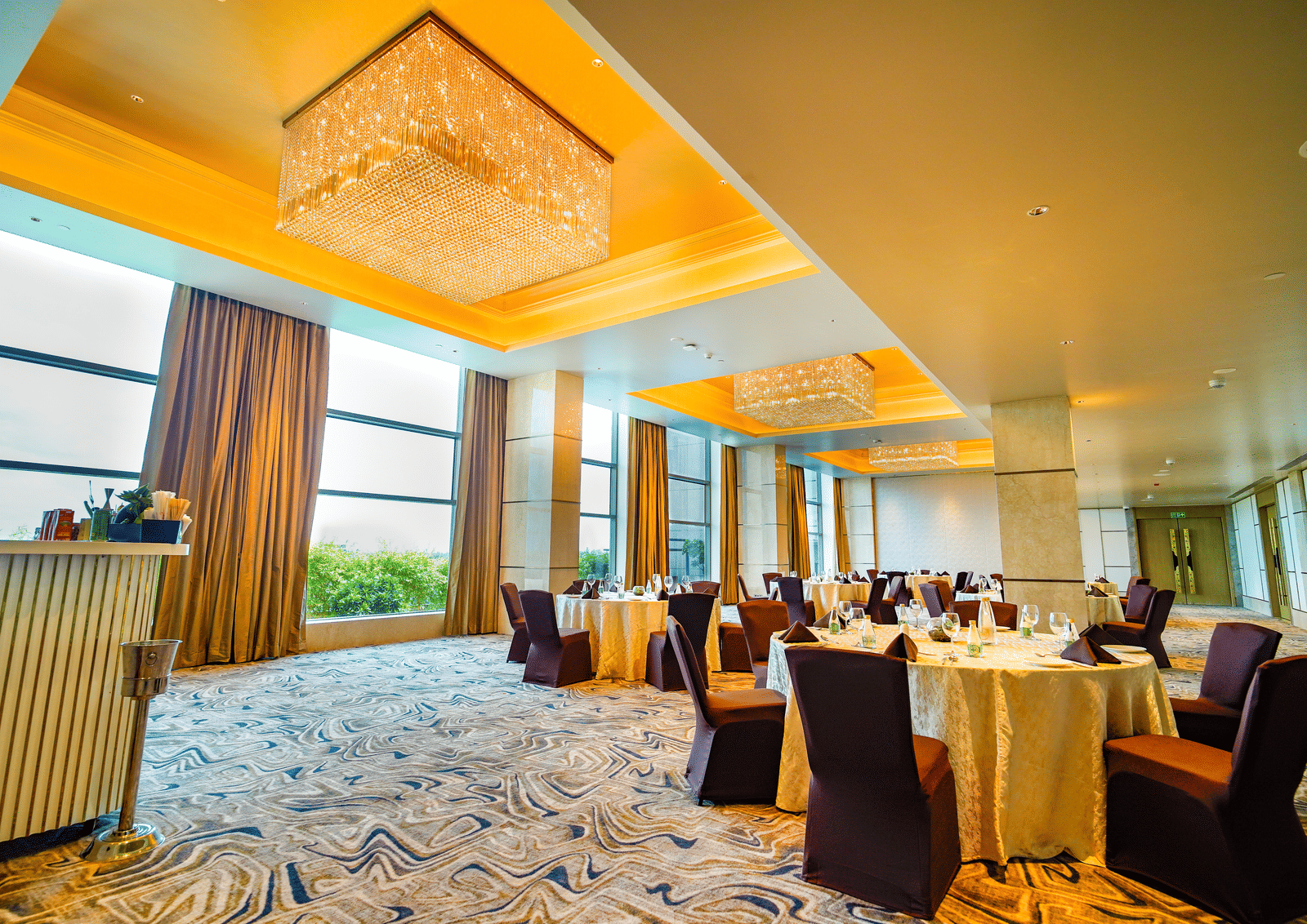A slightly lower angle shot of a  banquet hall with round tables, elegant chandeliers, and natural light from large windows - Grand Madurai by GRT Hotels