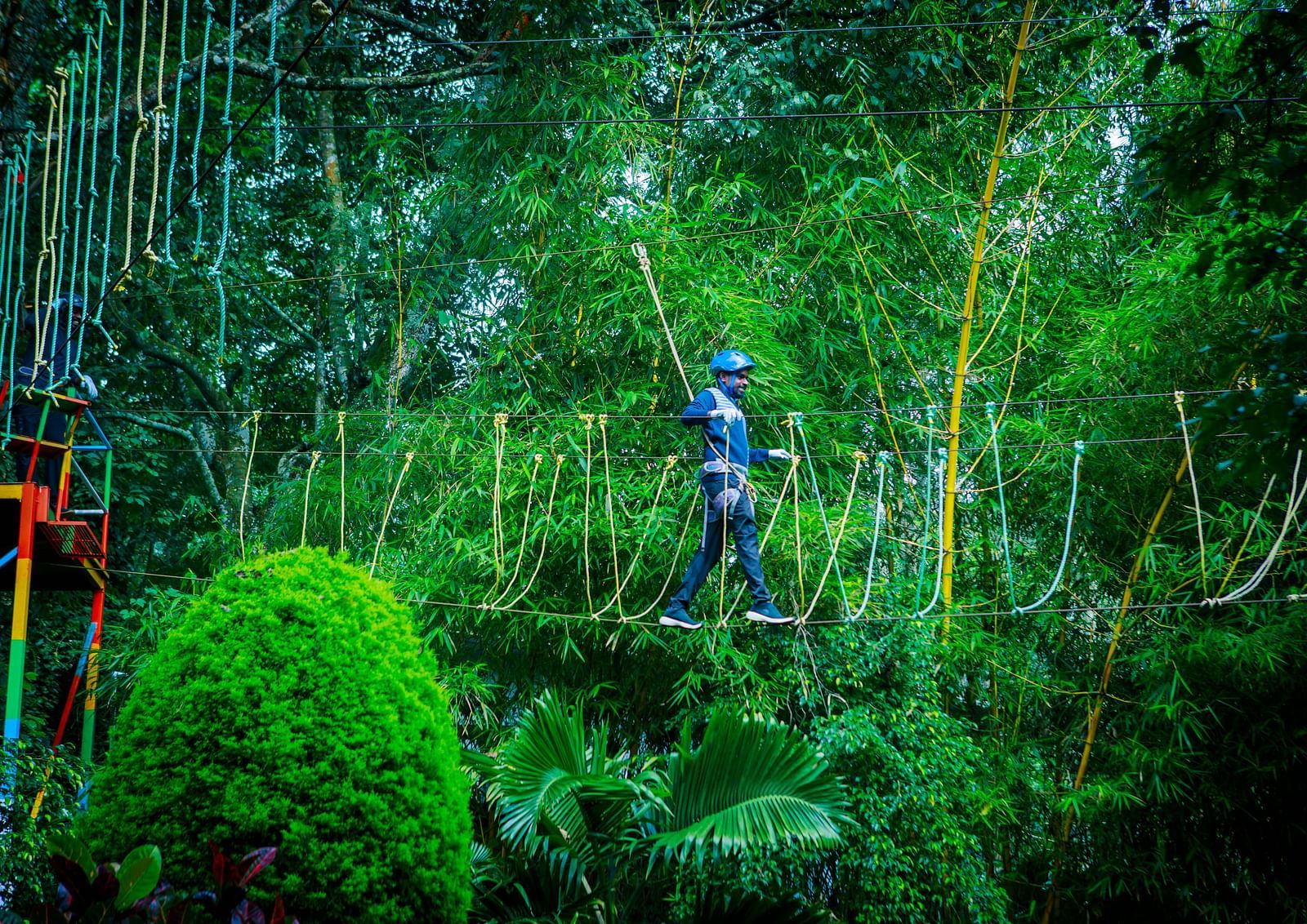 A person wearing a helmet and safety harness walks across a rope bridge in a lush forest canopy. - GReaT trails Yercaud by GRT Hotels