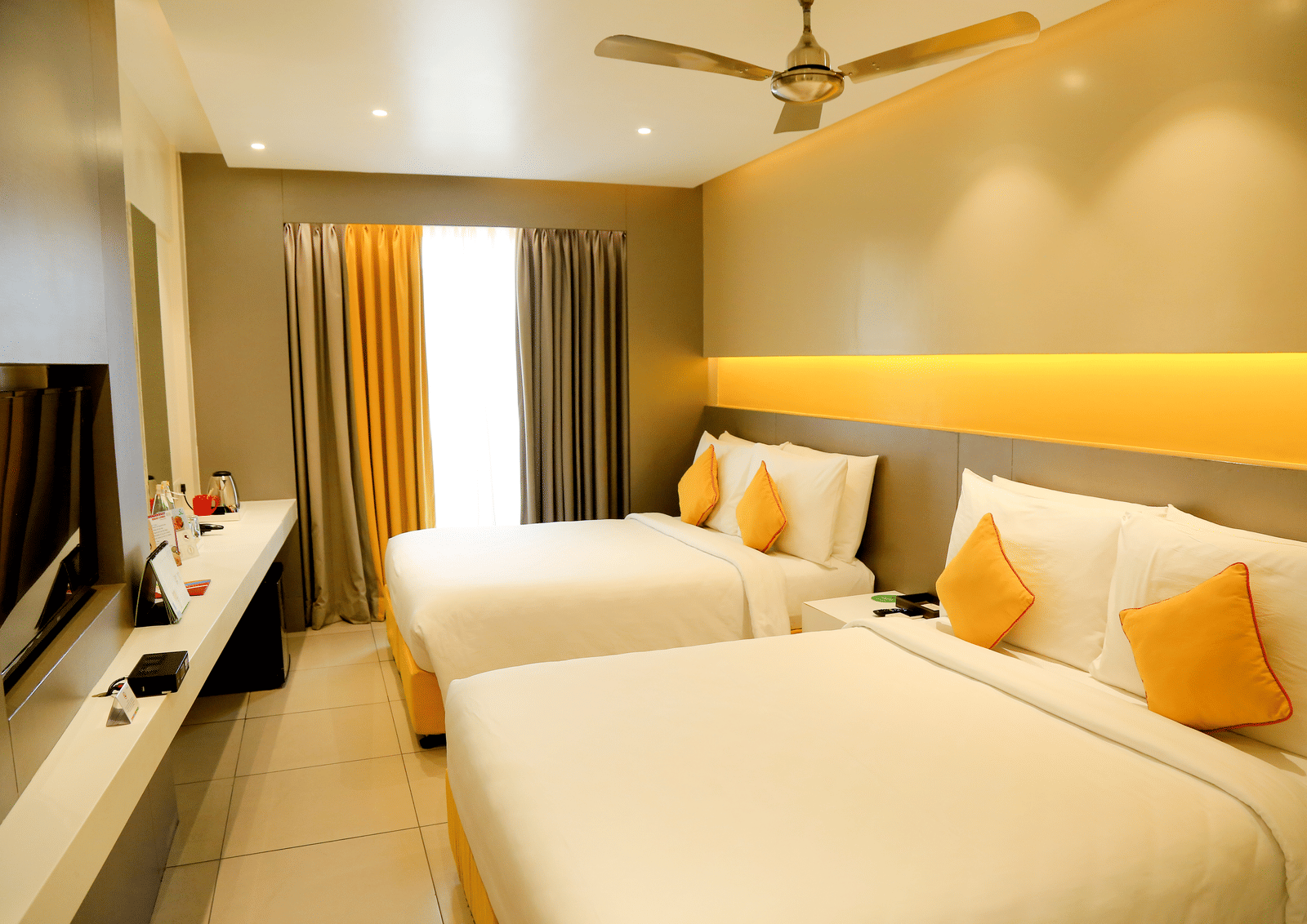 A spacious room with twin beds featuring yellow cushions and headboards, large windows with curtains, and a wall-mounted TV - Zibe Coimbatore by GRT Hotels