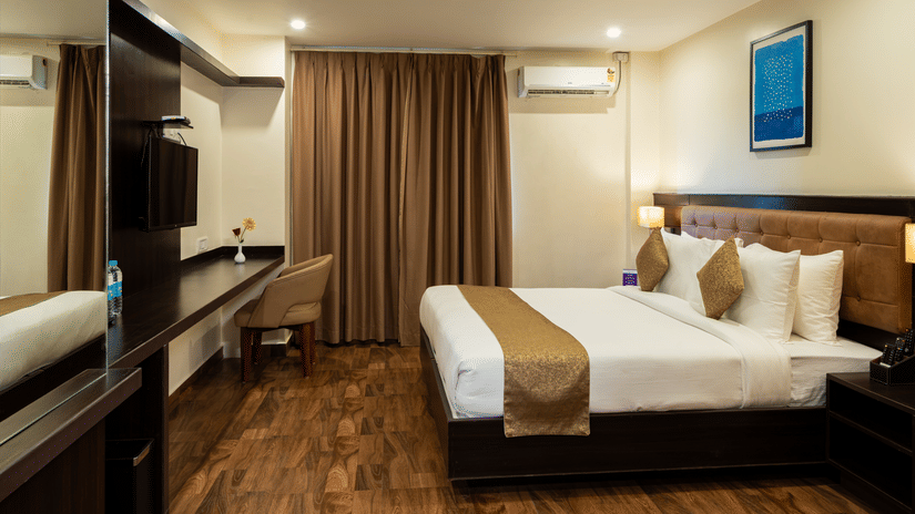 An elegant bedroom at VITS Select, Bengaluru, featuring a plush bed, rich wall paneling and curtains, a reflective mirrored surface, and a mounted air-conditioning unit for the perfect stay in Bengaluru.