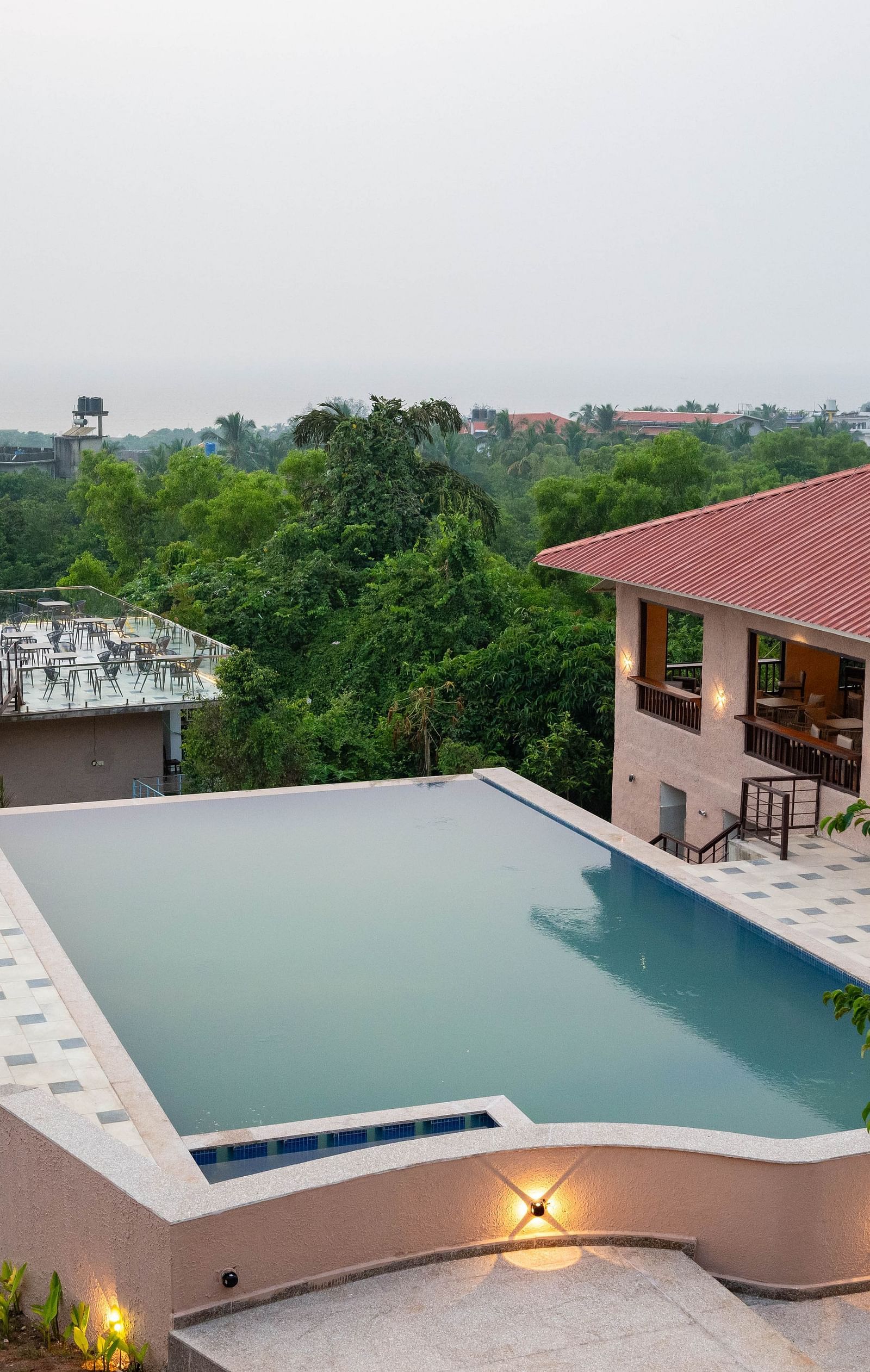 A designer swimming pool surrounded by bushes, with a small roofed building nestled beside lush greenery in the background. - Crescent Resort, Goa
