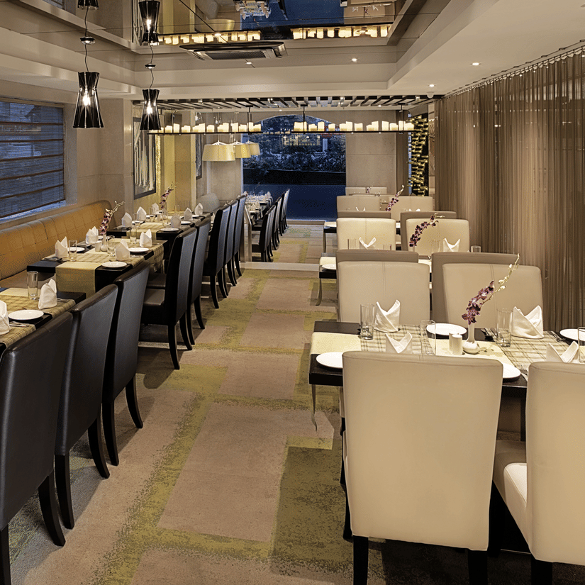 Interior view of Bahula restaurant at Barsana Boutique Hotel, Kolkata, showing organised rows of dining tables with neat place settings and contemporary decorative lighting.