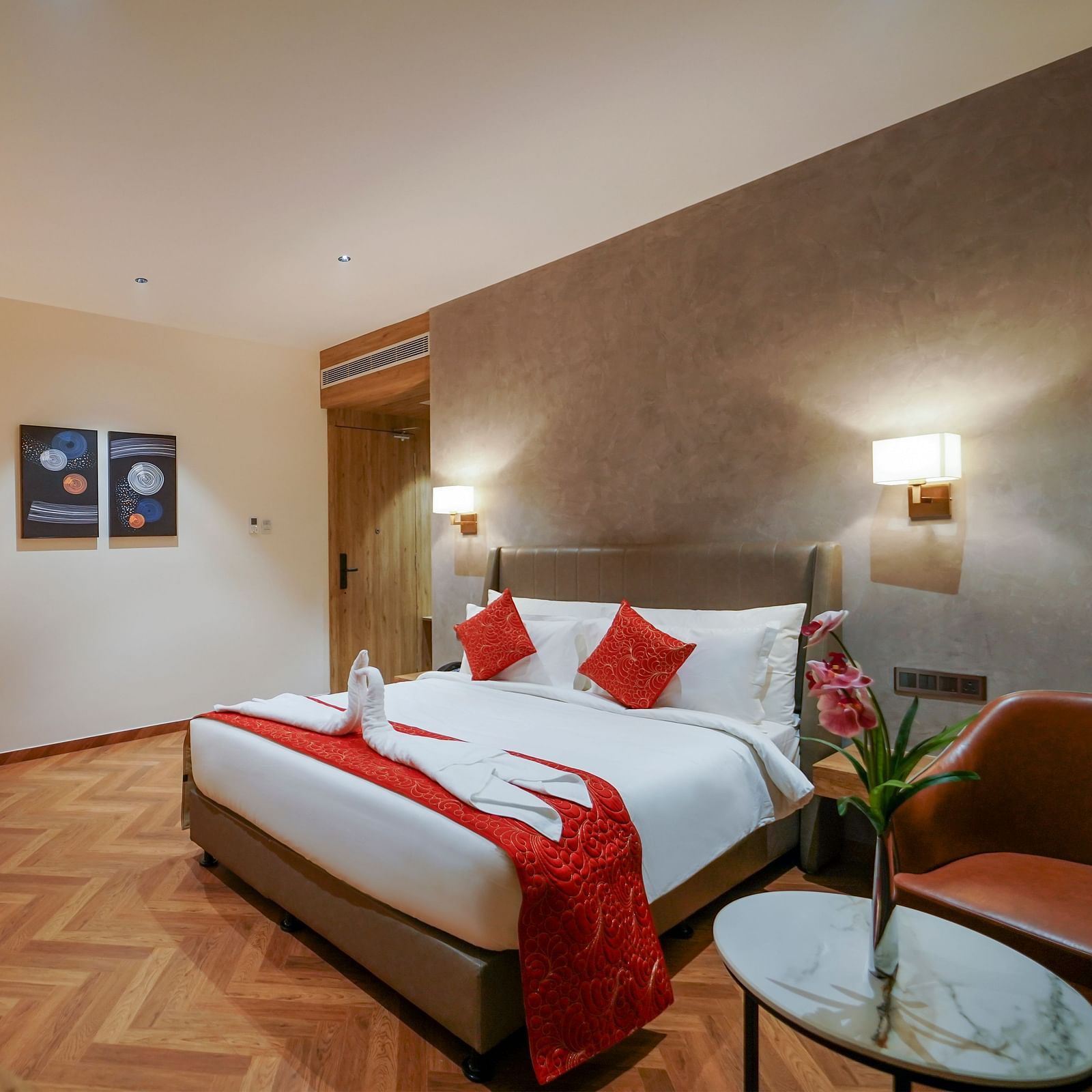 A hotel room includes a bed with pillows and folded linens, a desk area with a chair, a round table with flowers, and artwork on the wall - Hotel Park Inn, Nellore.