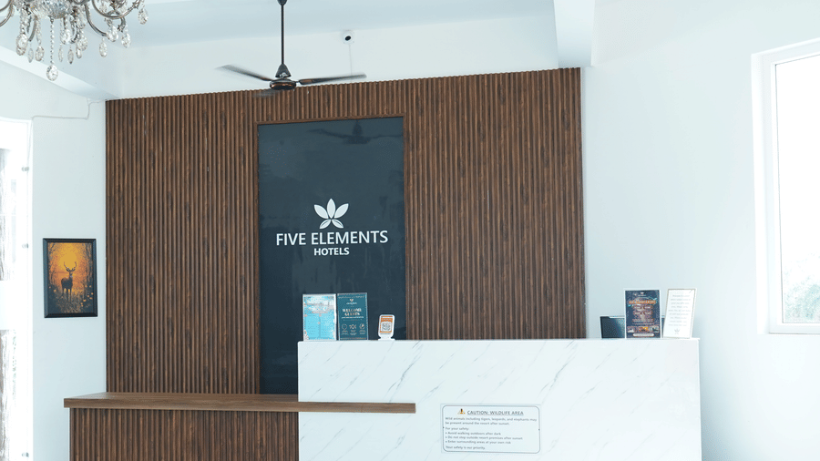 Reception area featuring a modern front desk counter with branding, wood paneling background, crystal chandelier, and bright tiled floors - Five Elements Hotels, Corbett Crown Resort, Jim Corbett