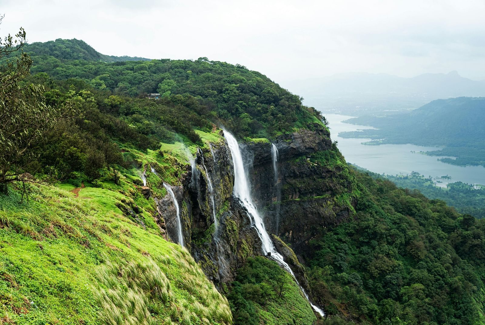A hillside in Matheran, Maharashtra, featuring a waterfall cascading down the rocks and lush greenery covering the landscape.