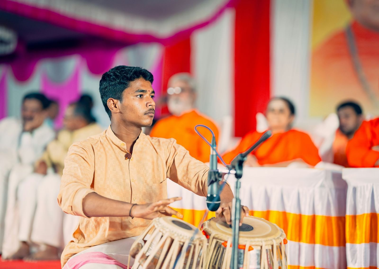 A person in a light-colored tunic is playing a pair of tablas on a stage with a microphone.