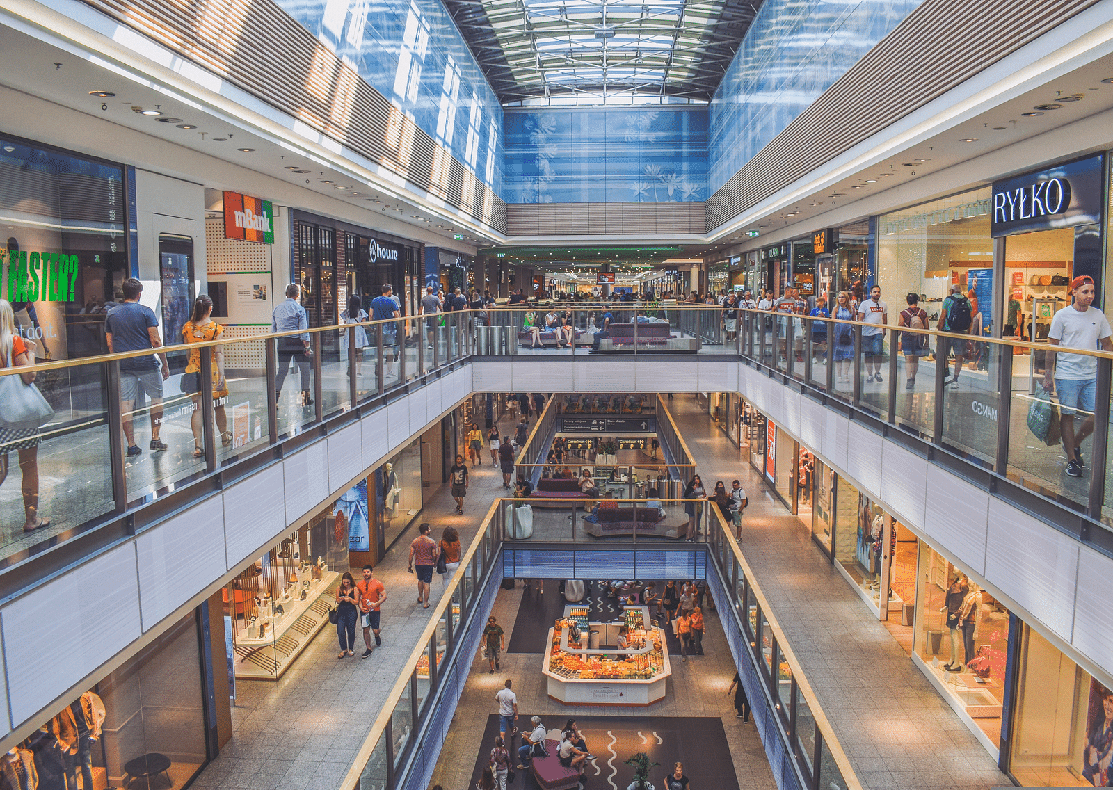 An overview of a mall with glass roof and a view of two floors with people walking.