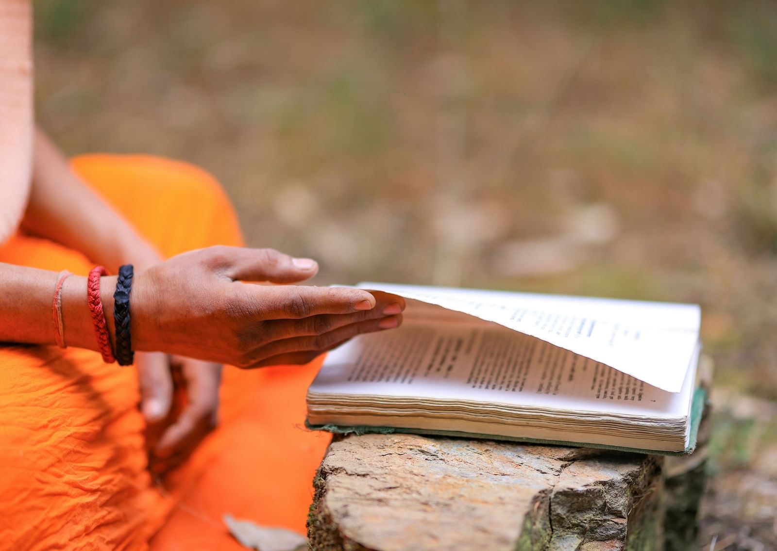  A close up shot of the hands of a monk in an orange robe turns a page in a book resting on a rock. 