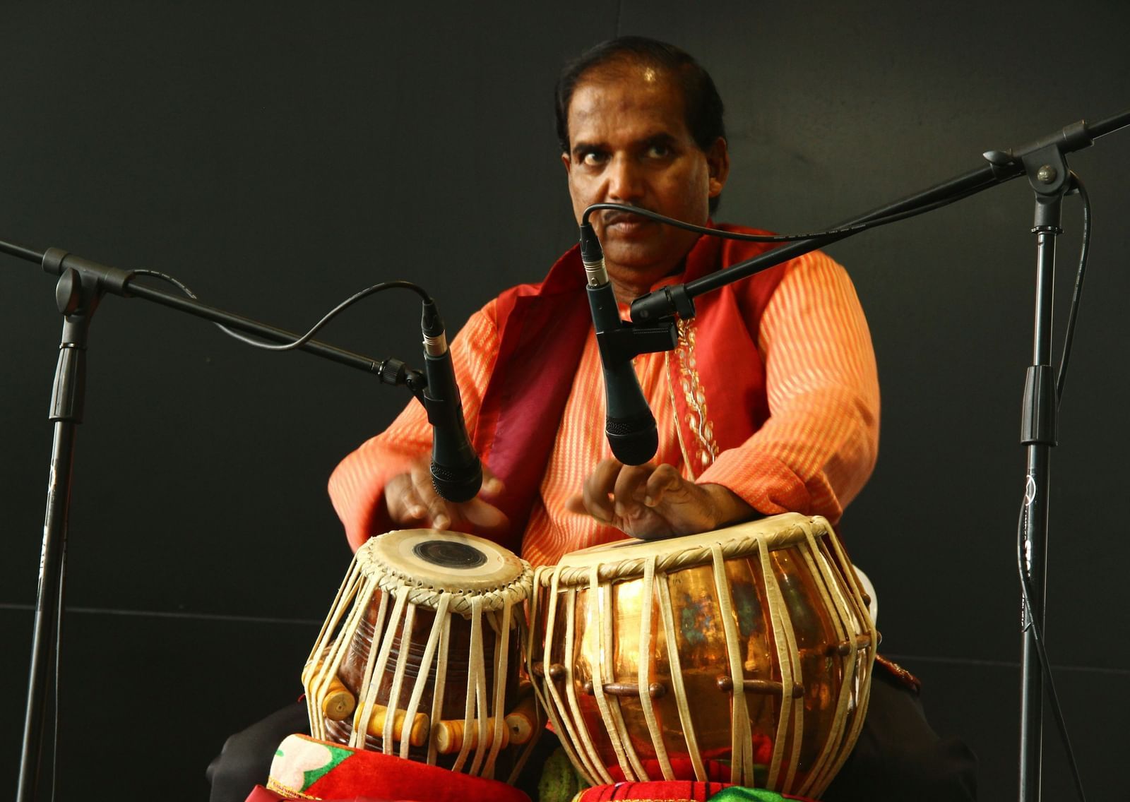 A man on a stage playing a pair of tablas, with microphones in the foreground.