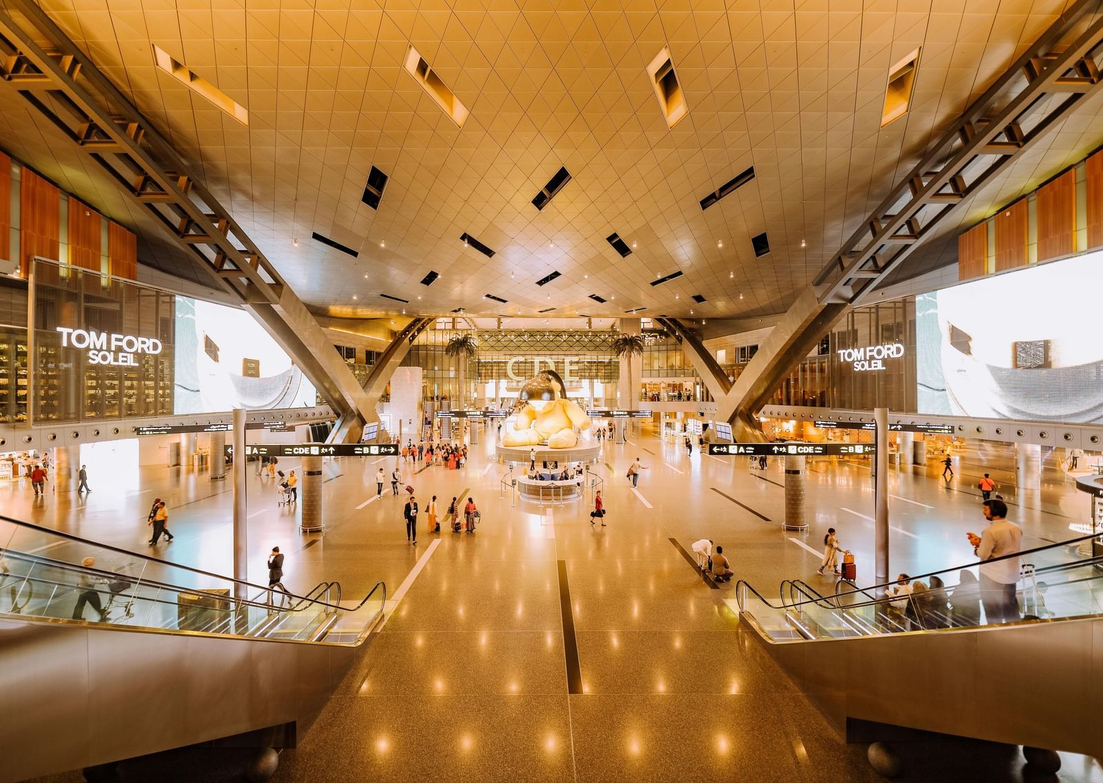 A grand, warmly lit shopping atrium featuring wide walkways, glowing ceiling lights, and elegant storefronts on multiple levels.