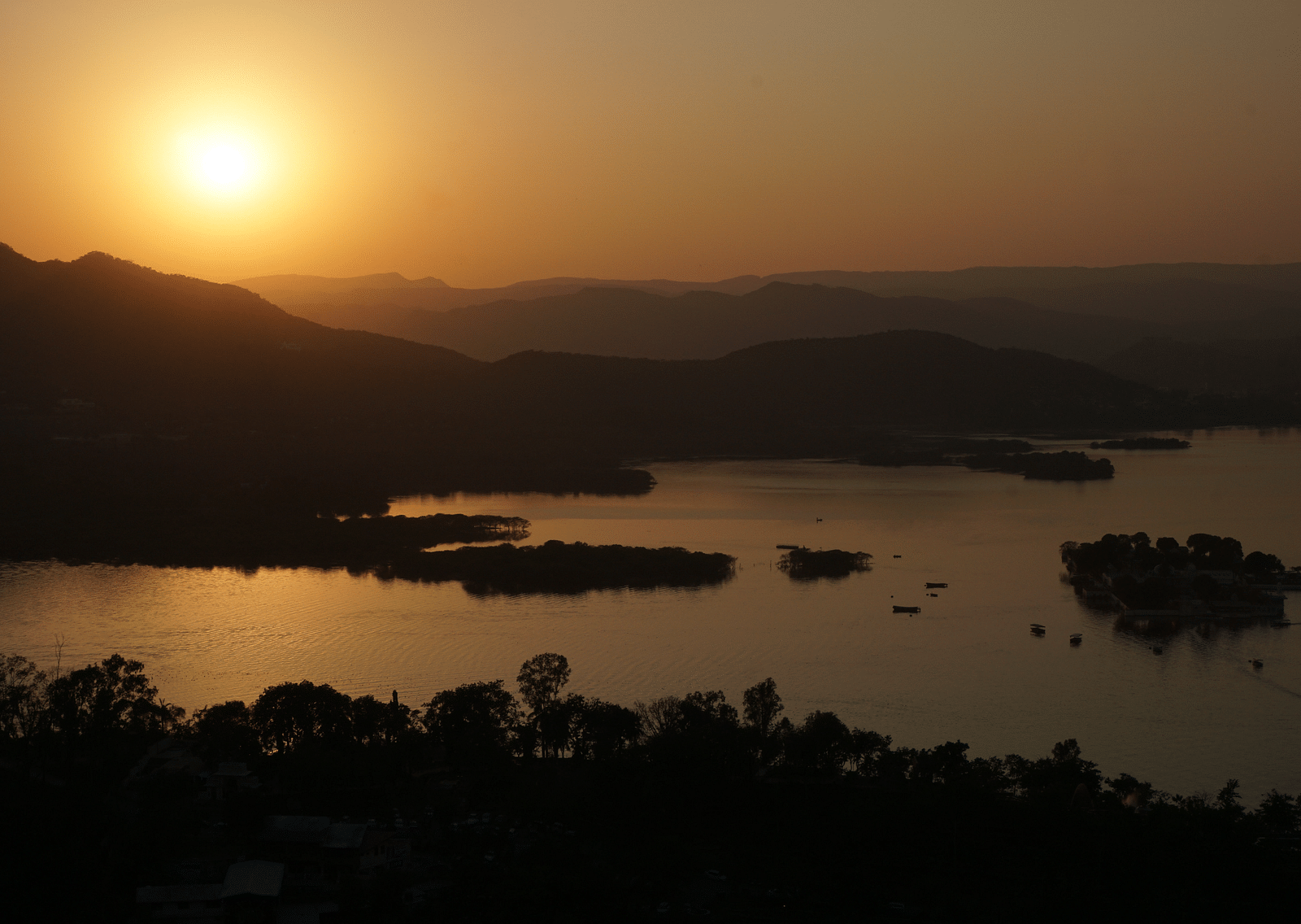 An overview of a sunset in lake pichola with hills and trees in view.