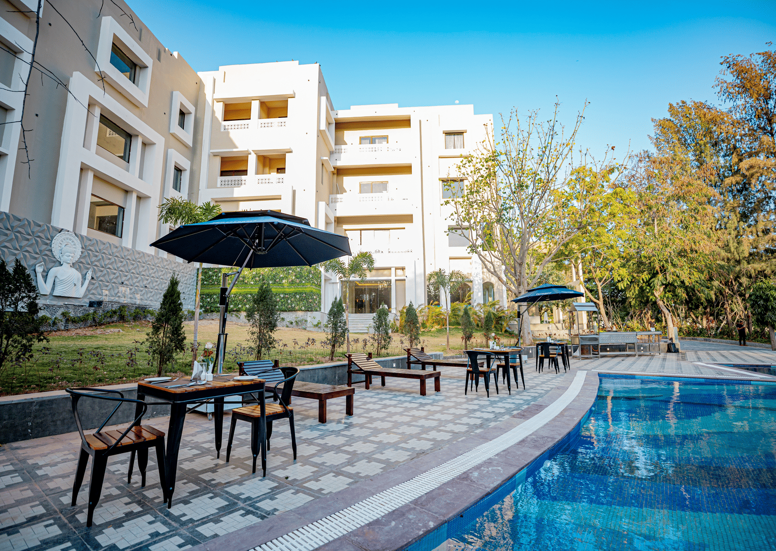 Dining setting placed by the pool with the facade of Essentia Luxury Resort & Spa, Udaipur in the background surrounded by trees