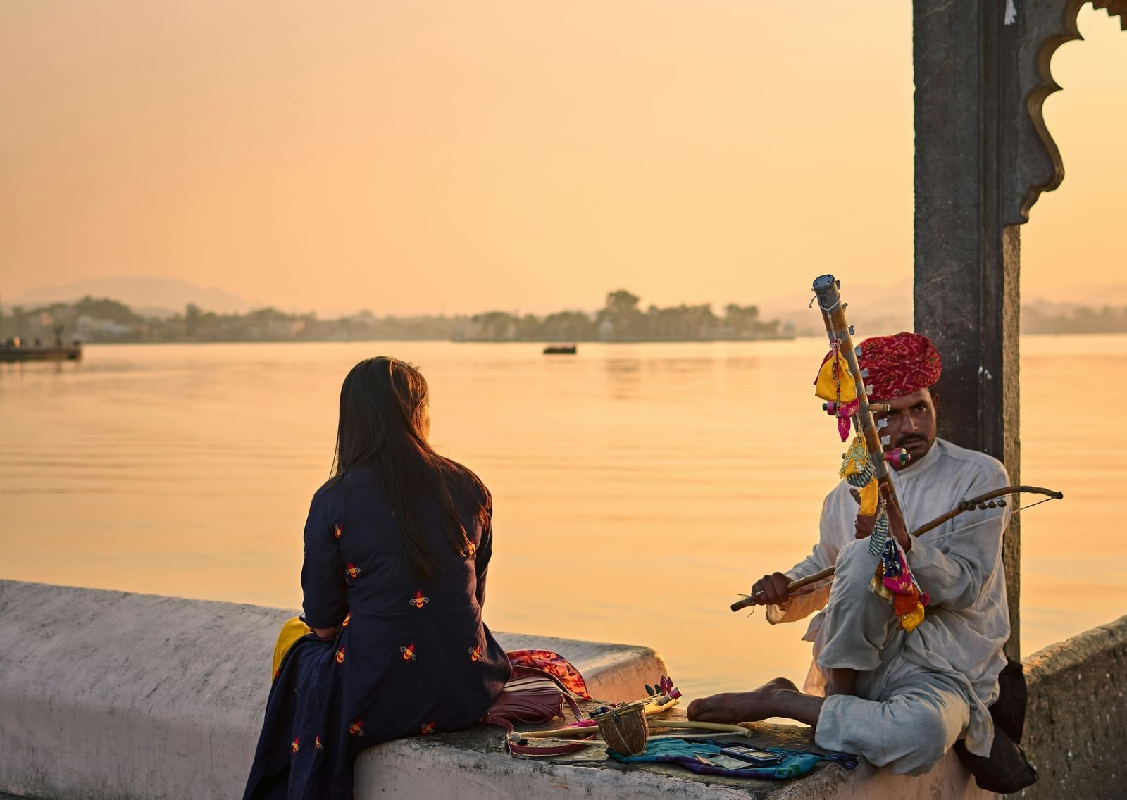 Person sitting by Lake Pichola at sunset with a traditional musician nearby.