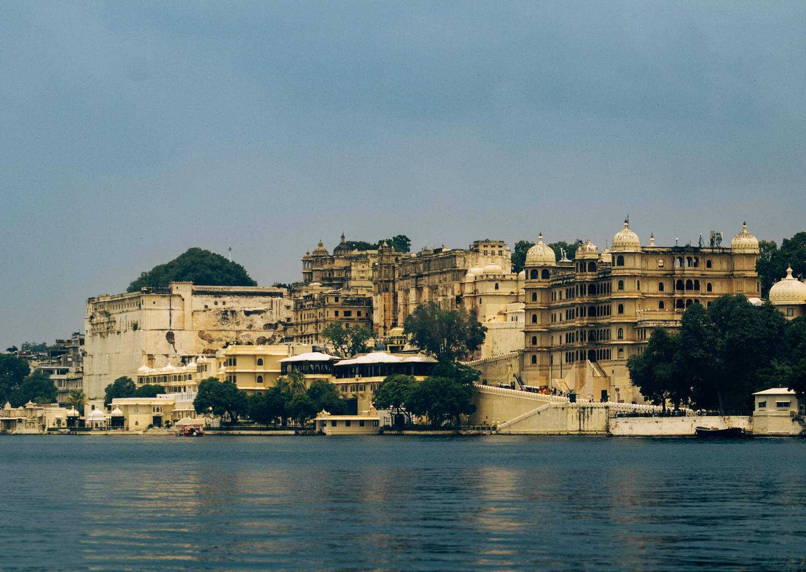 View of the City Palace complex in Udaipur, Rajasthan, reflected in the calm waters of Lake Pichola under a clear sky.