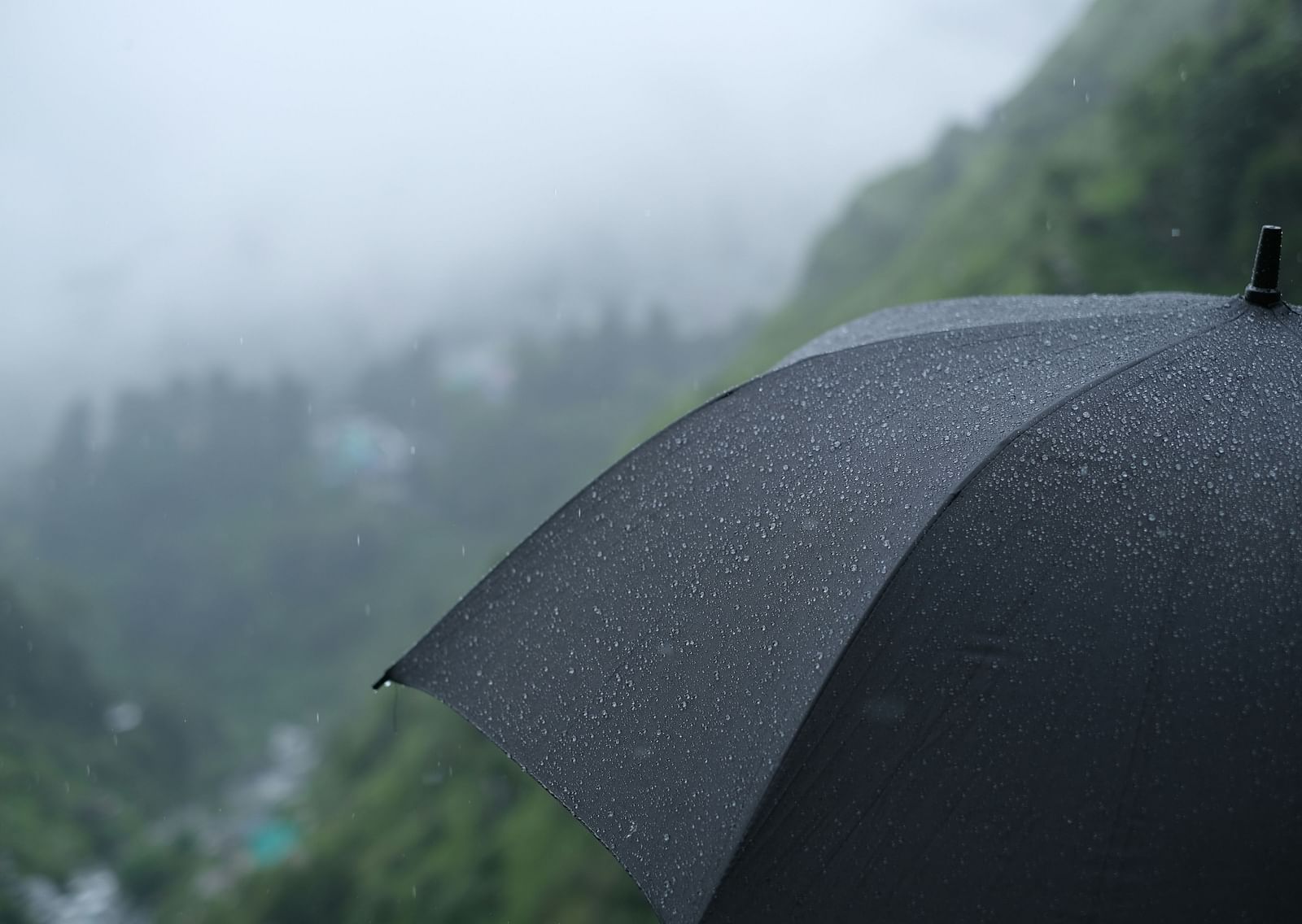 A black umbrella covered in raindrops, set against a misty, green mountainous landscape during a gentle rainfall.
