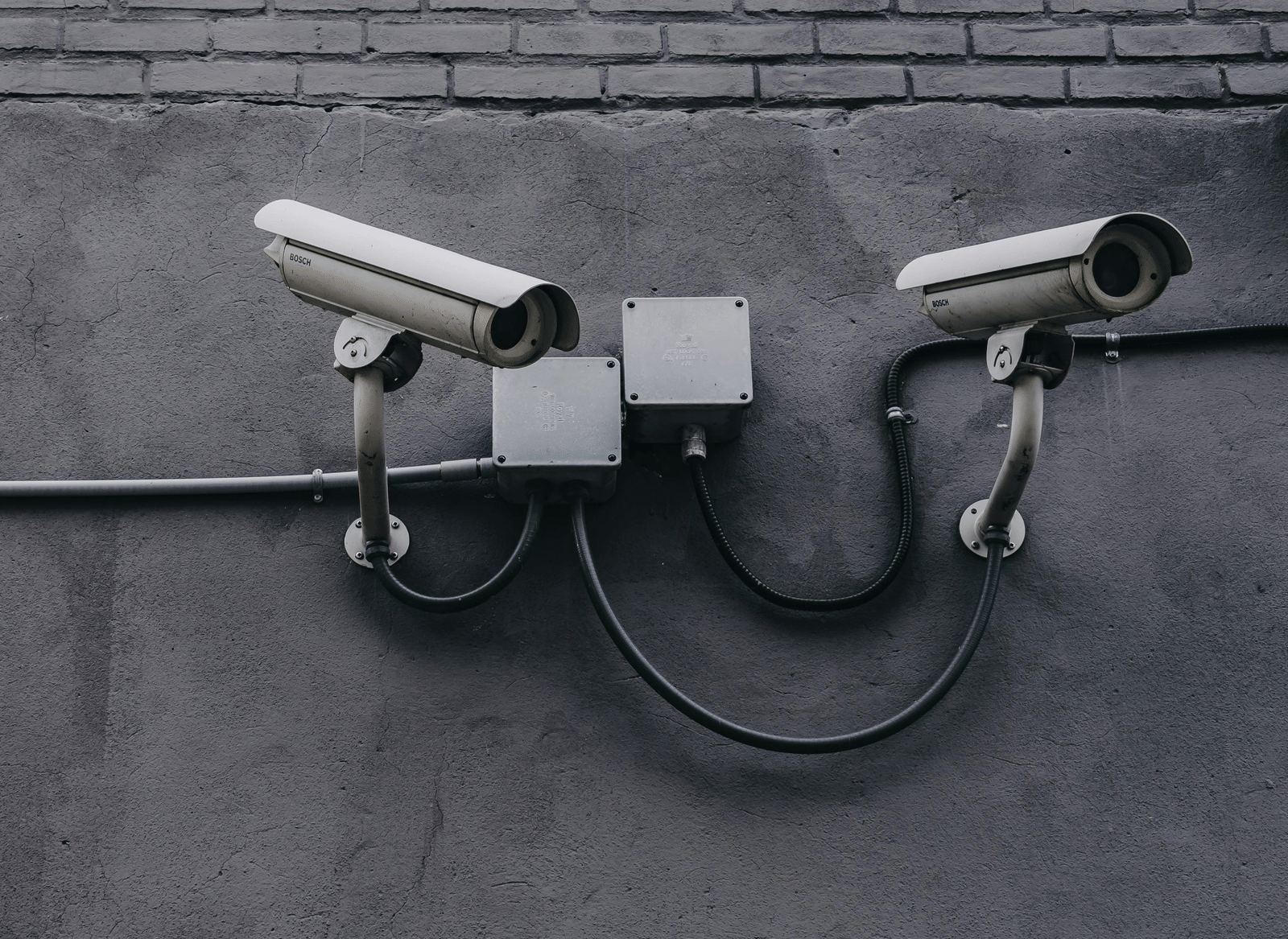 Two surveillance cameras mounted on a grey concrete or brick wall, with visible wiring.