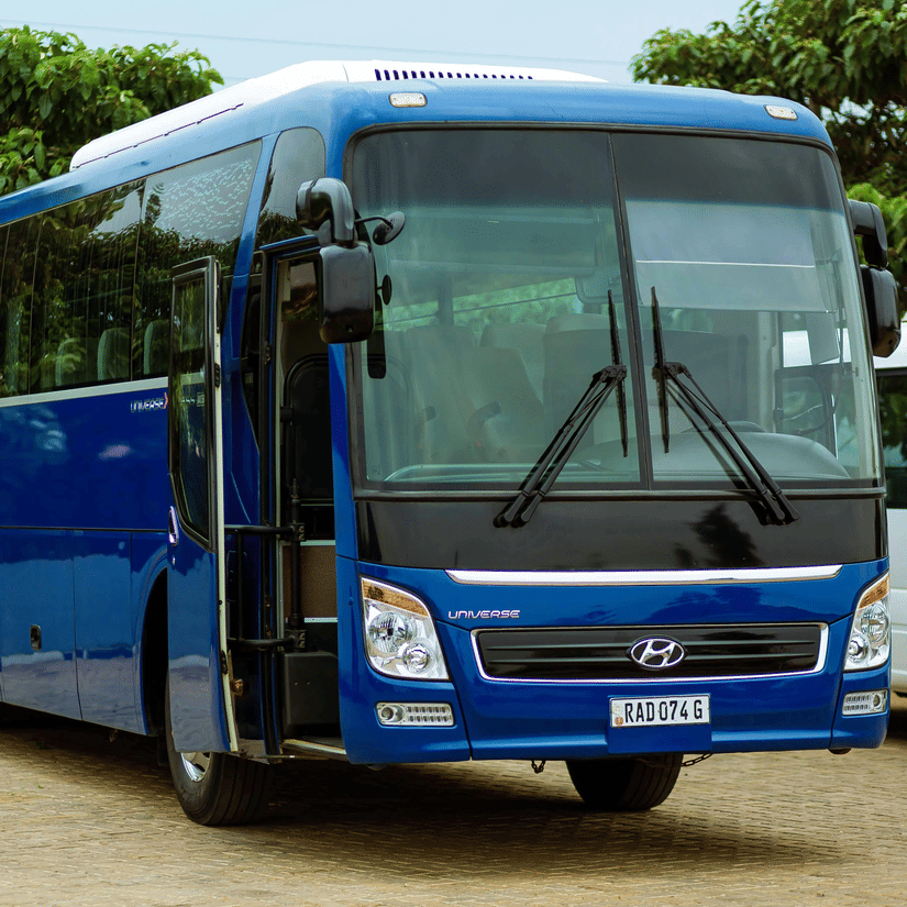 A blue coloured Hyundai bus, parked on a cobbled road, with another vehicle parked next to it and trees behind it.