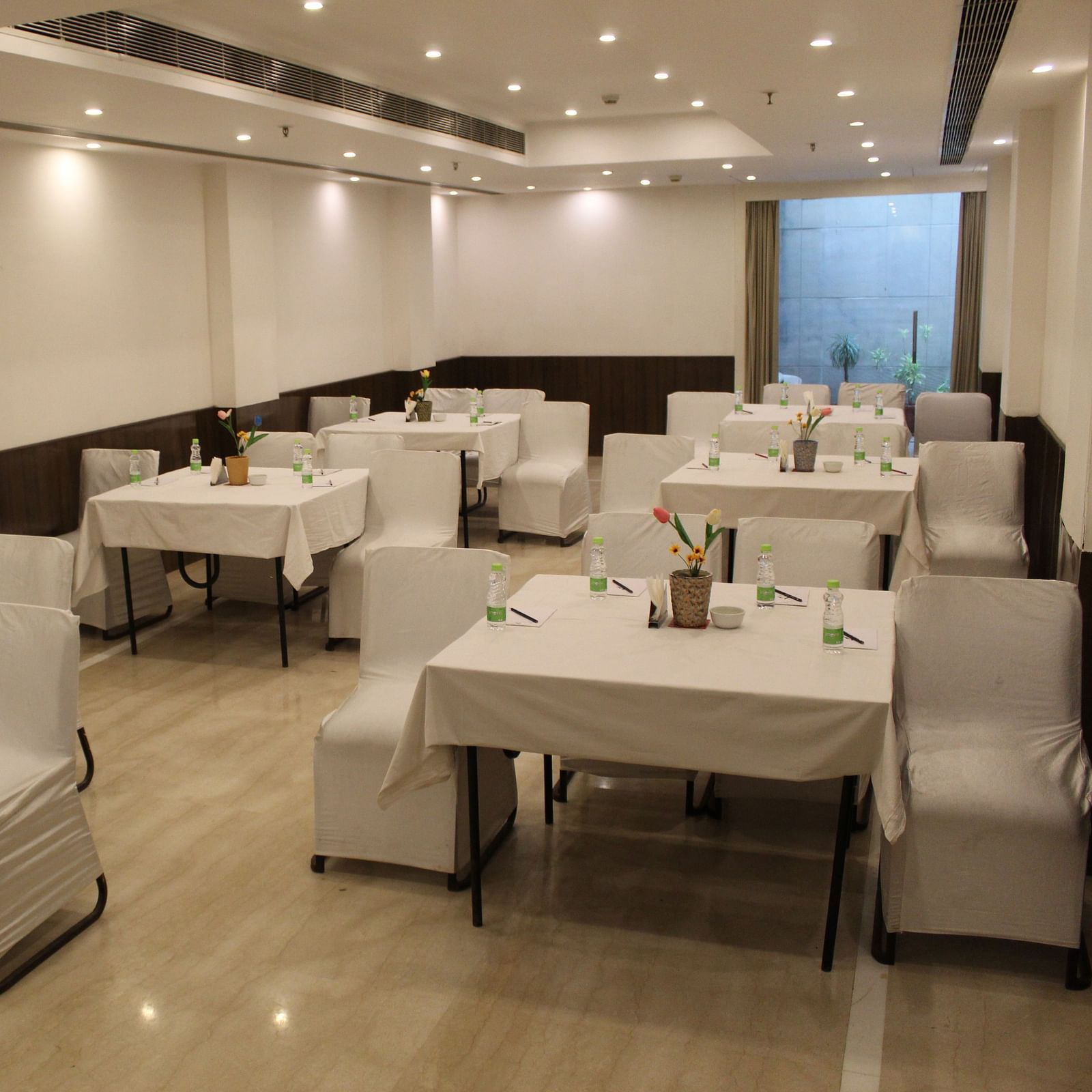 An image of a banquet hall at Inde Hotels Vista Woods featuring several sets of chairs and table where water bottles and flower pots are placed 