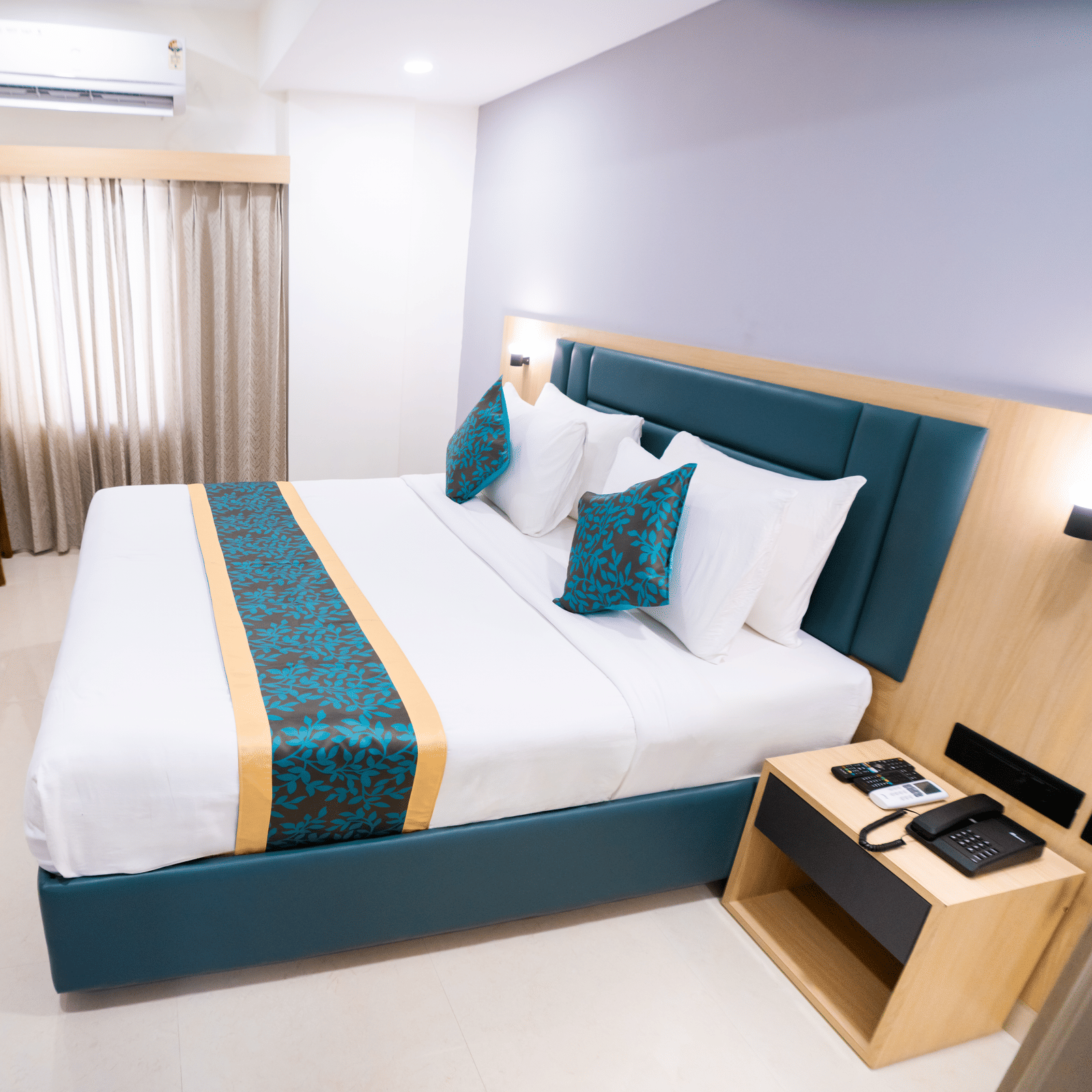 A side view of the room with a large bed, bedside table, wall-mounted TV, and a window with curtains at Sunrise Business Hotel, Hyderabad.