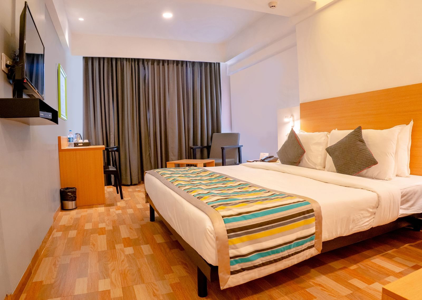 Another view of a cosy hotel room with a large bed, colourful throw and wooden furniture, including a desk with a TV above it - TGI Ameya Grand, Goa